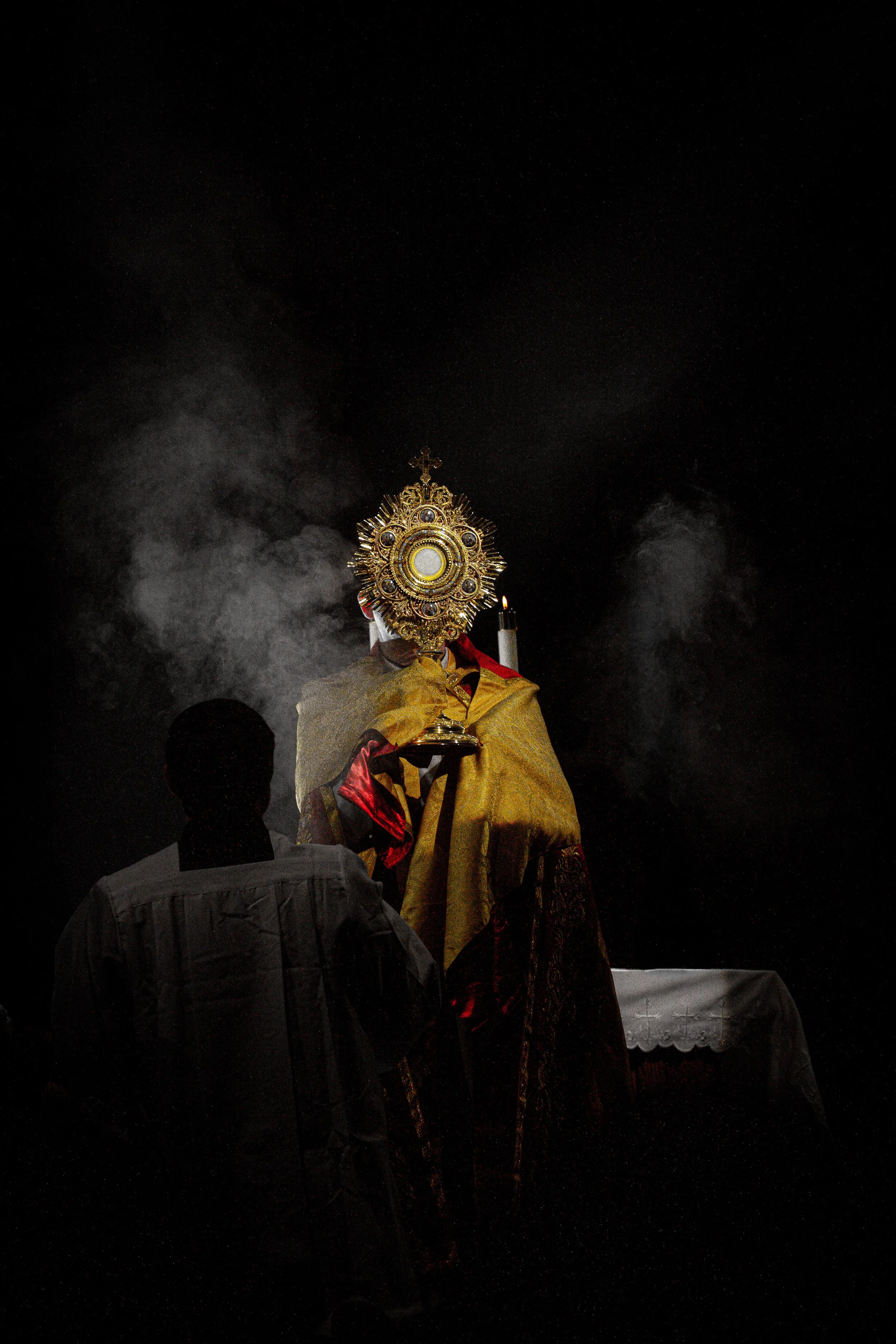 A priest dressed in ornate gold and red robes holding a large, decorative monstrance during a nighttime Catholic pilgrimage or procession with onlookers and incense smoke in the background.