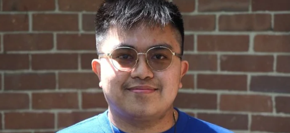 A young man with glasses and short dark hair, smiling, standing in front of a brick wall.