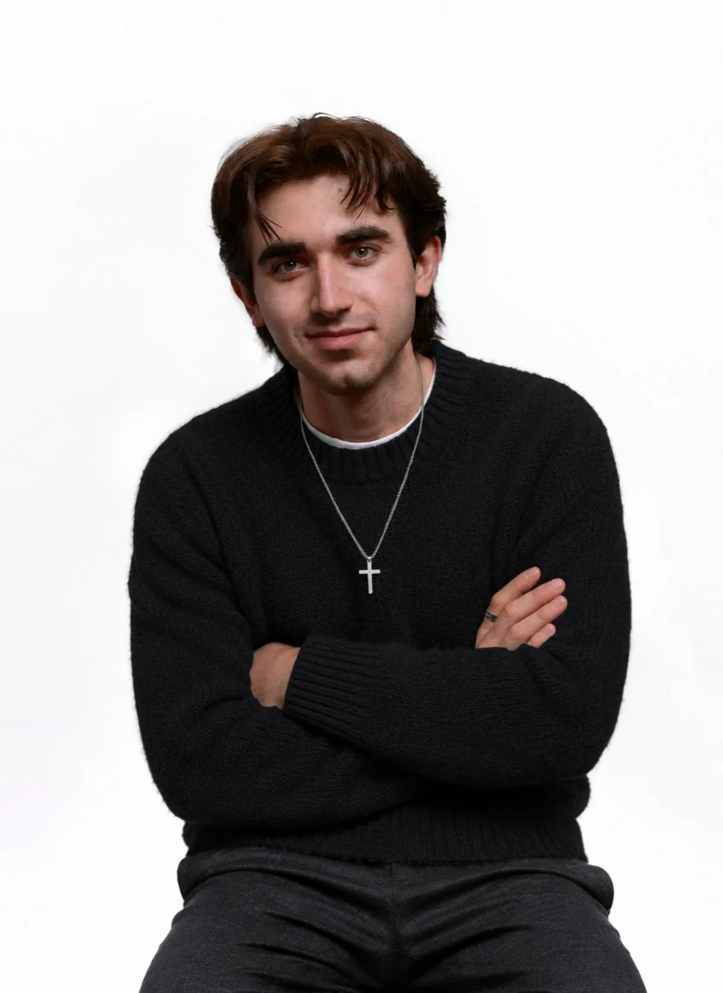 A young man with brown hair and green eyes wearing a black sweater, cross necklace, and dark pants poses with arms crossed against a plain white background.