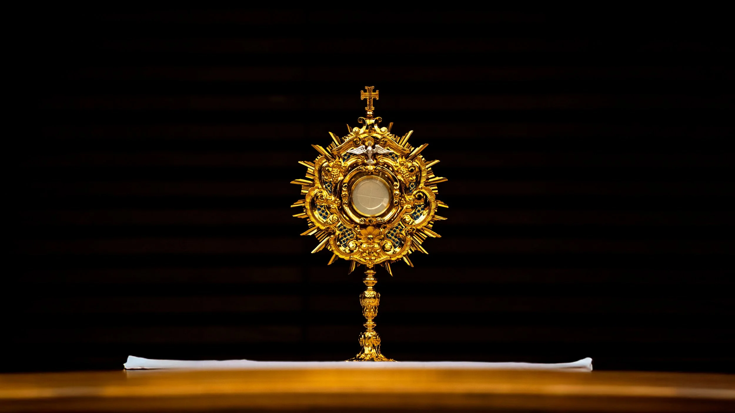 A gold monstrance with intricate details and a cross at the top, placed on a white cloth on a wooden surface, against a dark background.
