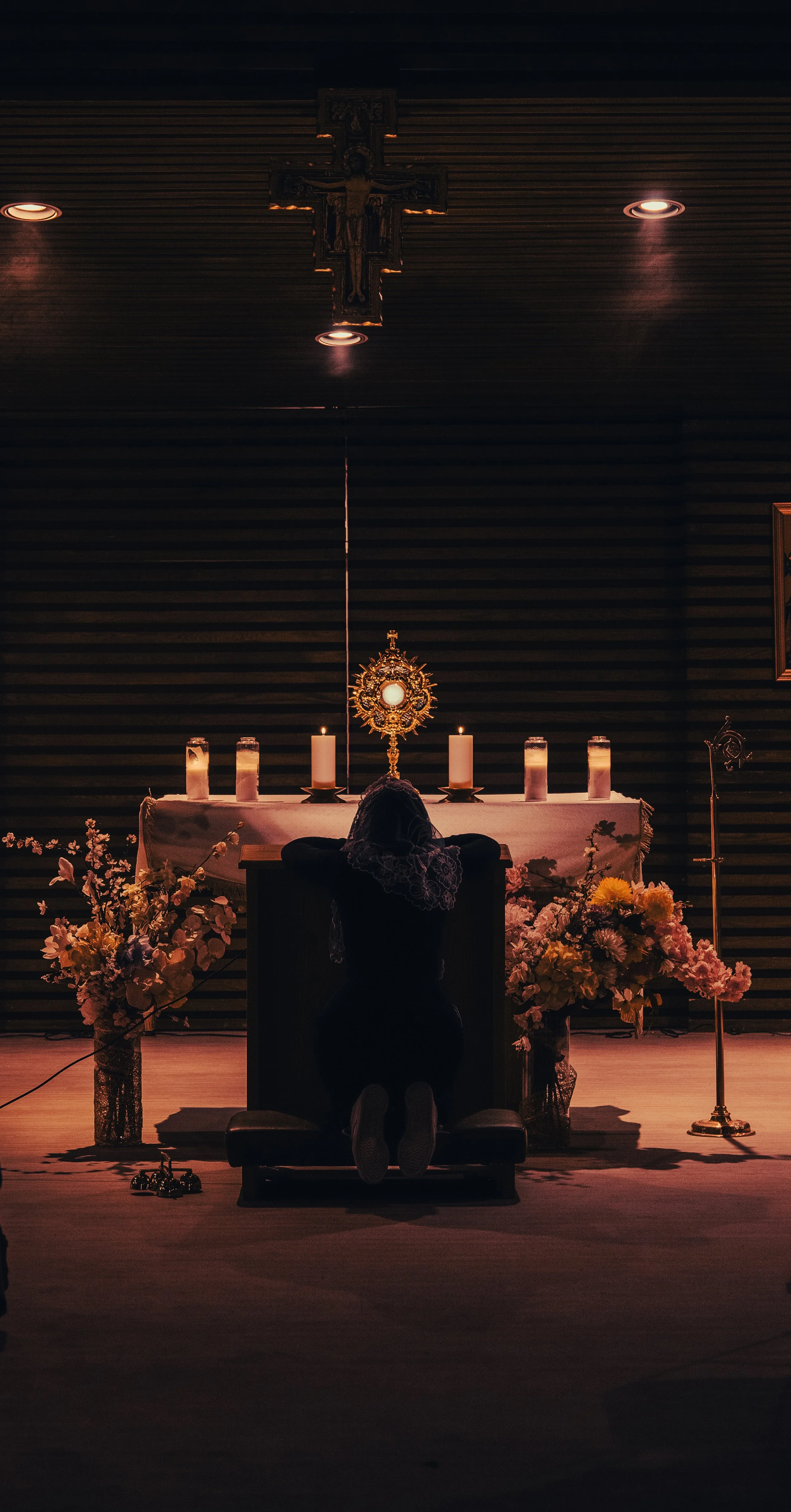 A person kneeling by a prayer altar with candles, flowers, and Christian religious symbols, including a crucifix and monstrance, in a dimly lit setting.