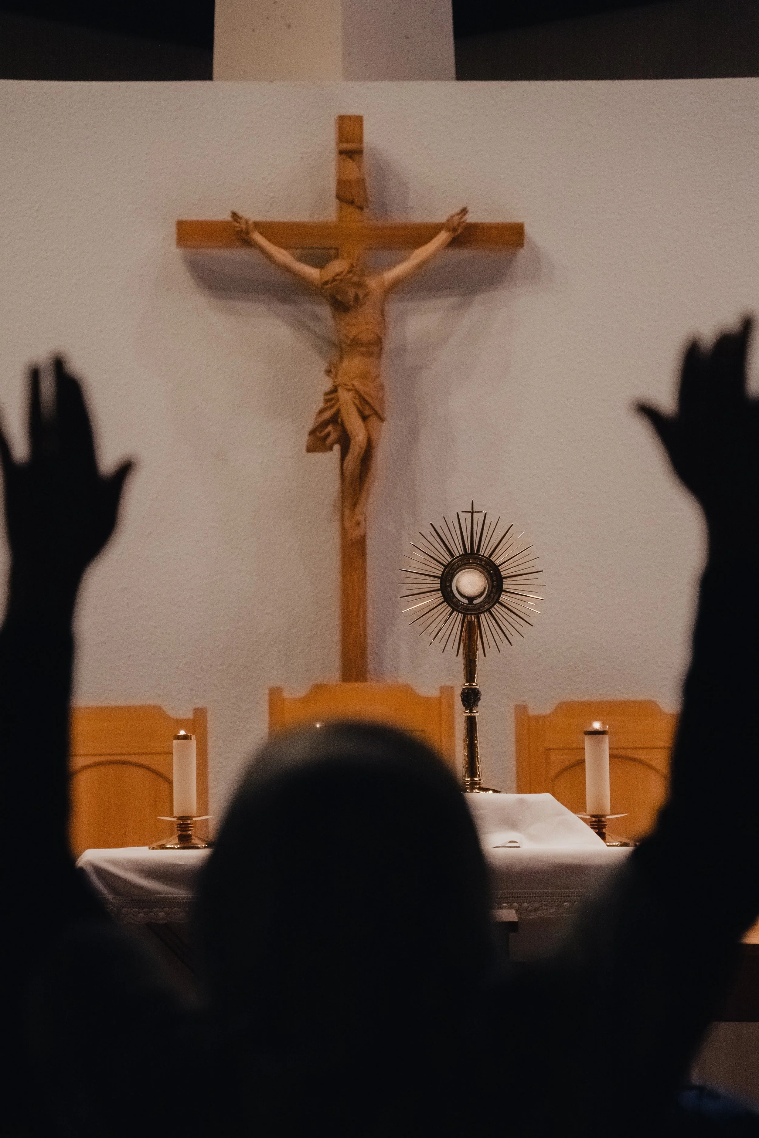 A wooden crucifix depicting Jesus Christ on the cross is mounted on a white wall. The altar in front of it has lit candles and a monstrance with a circular design and radiating spokes.