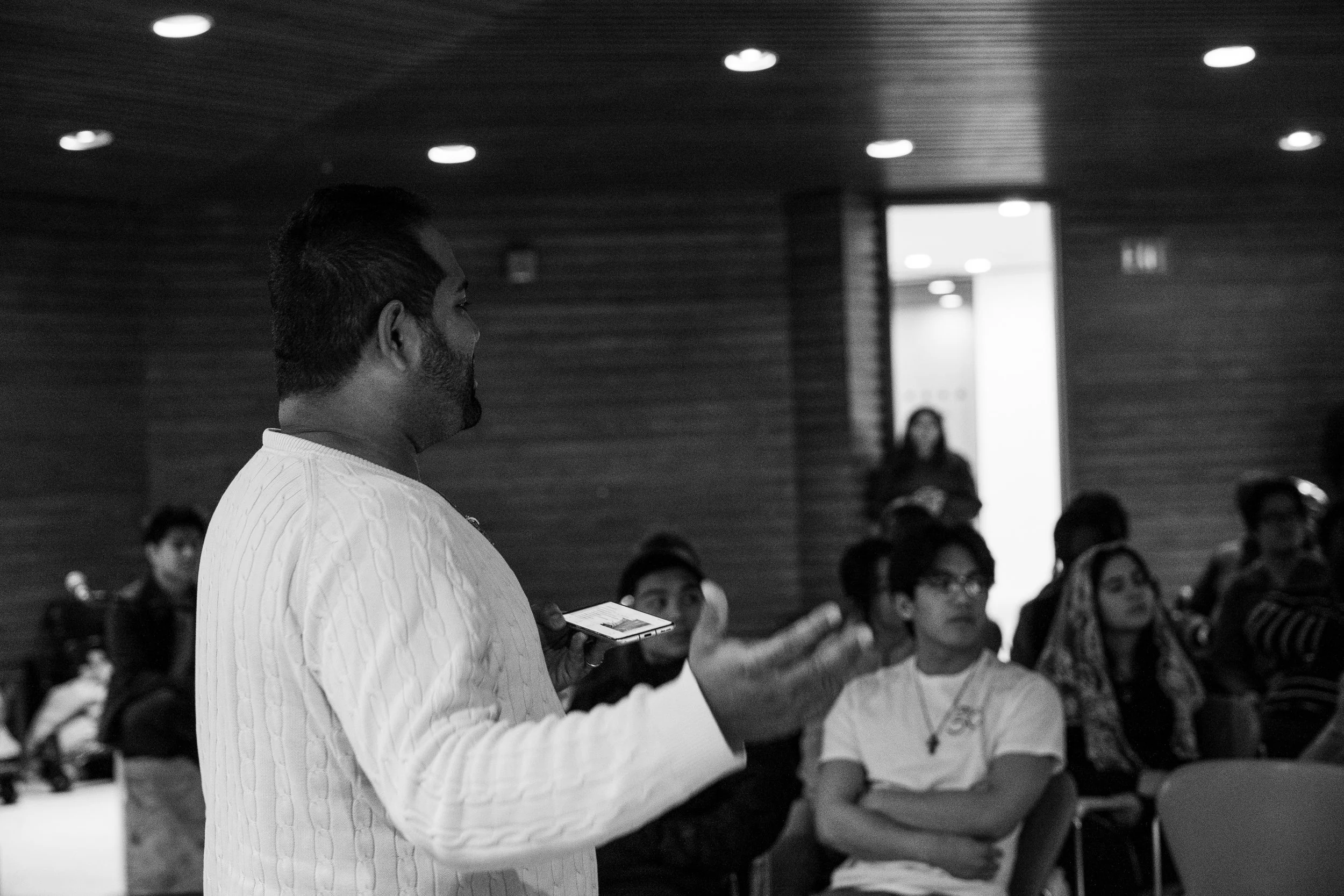 A man with dark hair and a beard wearing a white cable-knit sweater is standing and holding a smartphone. Behind him, a group of people, mostly women, are seated and appear to be listening attentively. The setting is indoors with wooden walls and ceiling, and overhead lights.