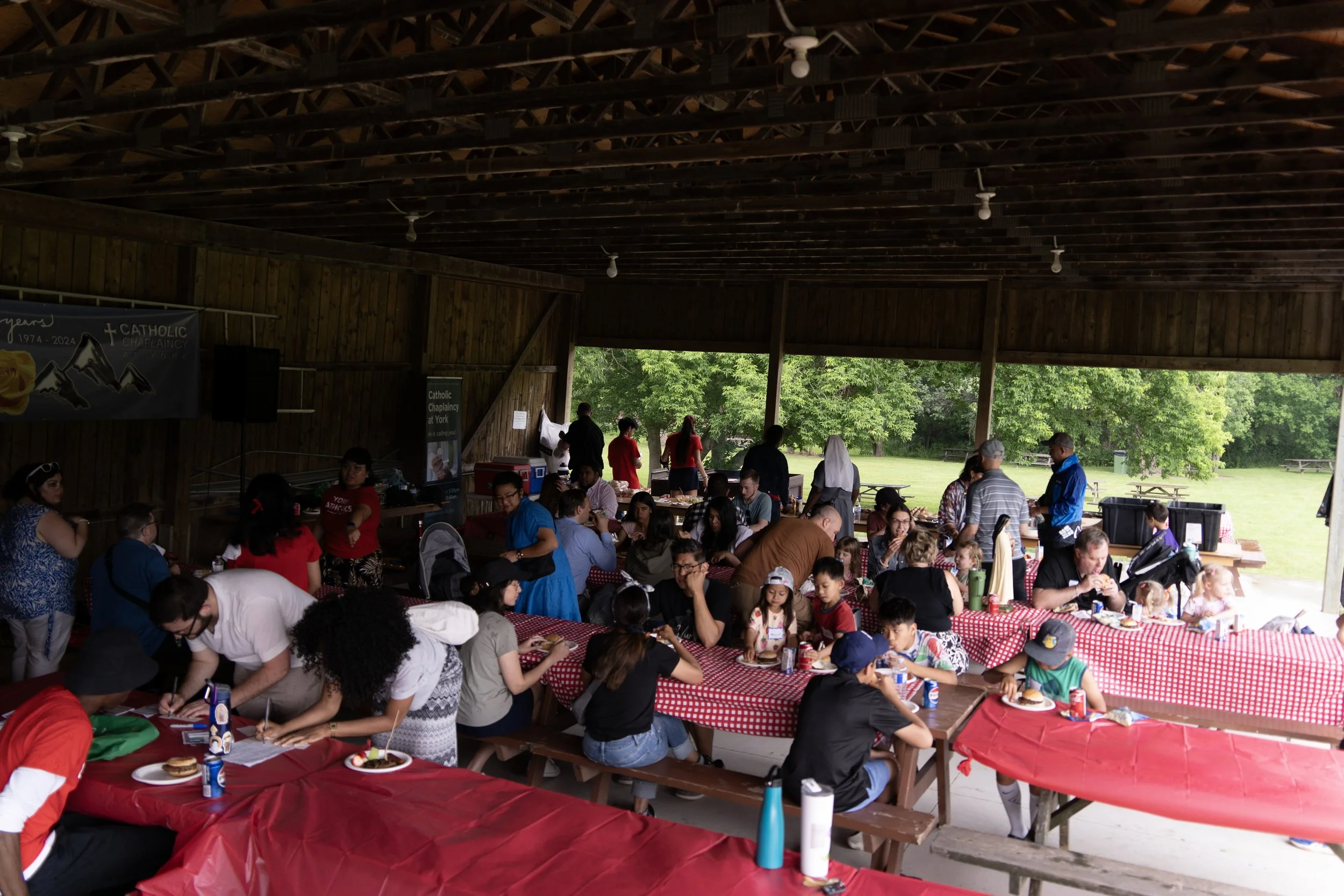 A large group of people gathers under a wooden pavilion during a community event, seated at long tables covered with red and white checkered tablecloths, eating and socializing. In the background, there are trees and open grassy fields, and some people are standing and serving food.