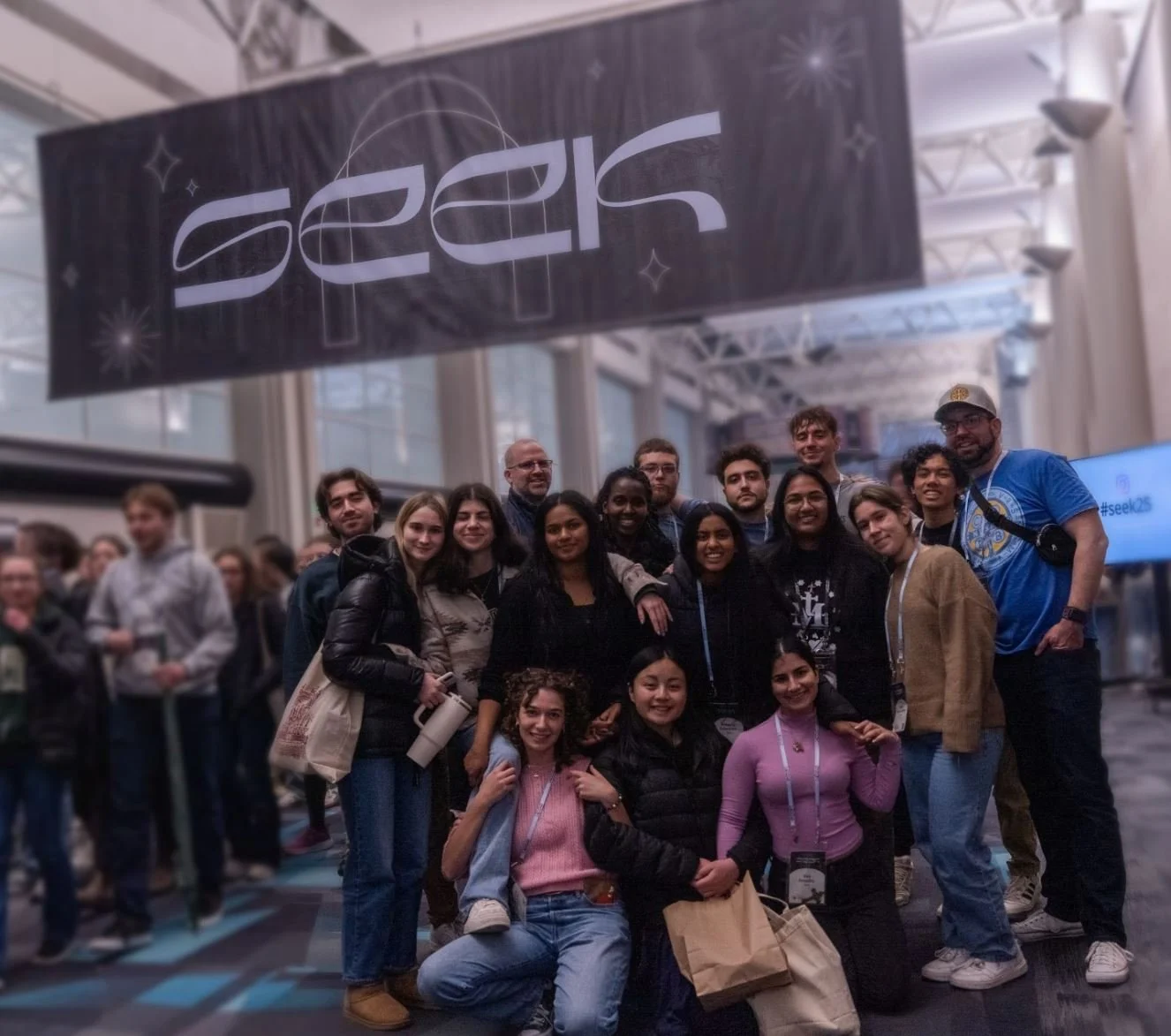 Group of diverse people posing at the SEEK 2023 conference, with a large SEEK 2023 banner overhead in a modern, well-lit venue.