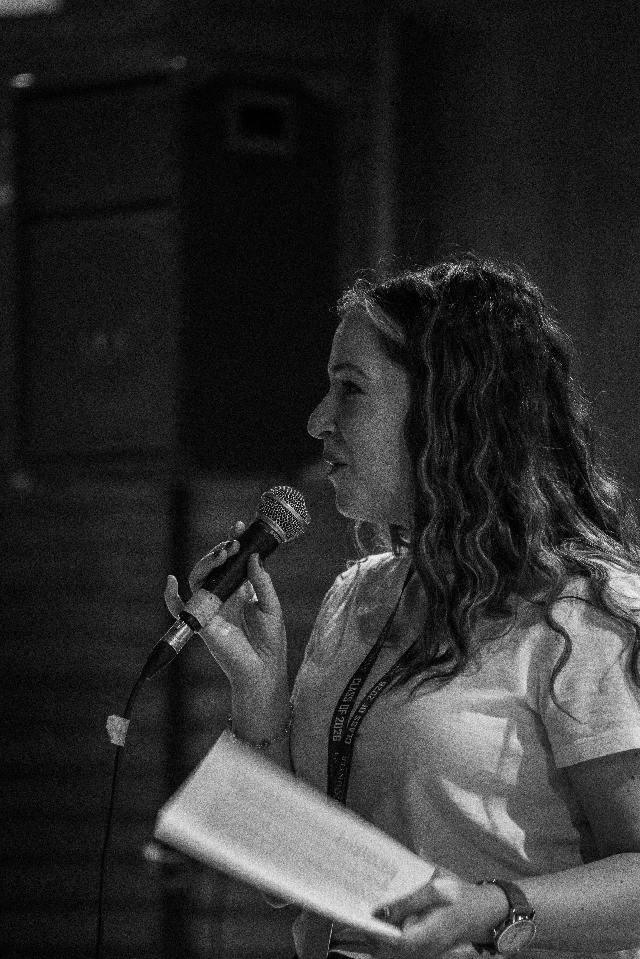 A young woman with long curly hair speaking into a microphone, holding a sheet of paper, wearing a bracelet and a watch, at an indoor event or conference.