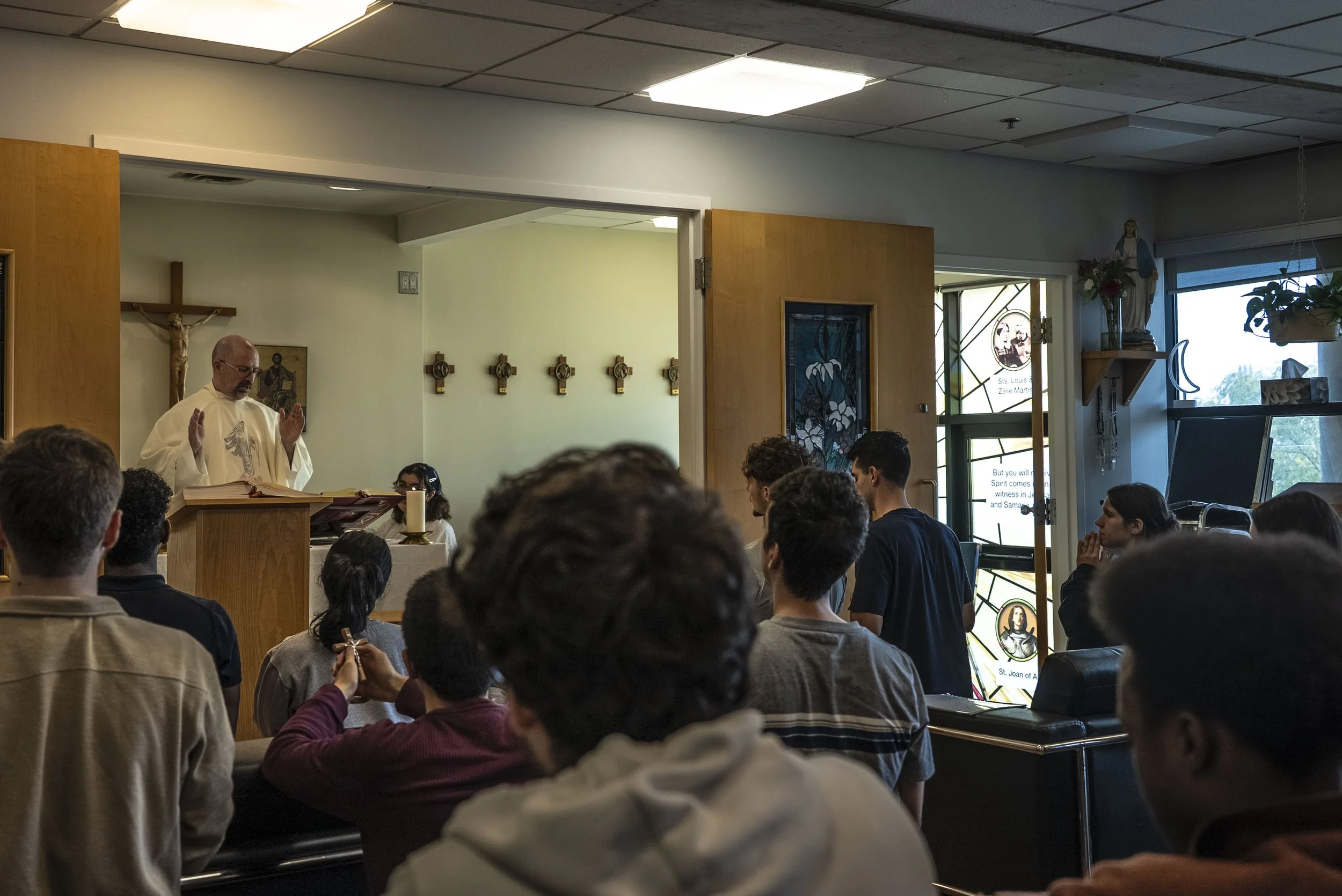 A priest standing at a lectern preparing to lead a religious service, with a congregation seated in a church or chapel interior. The priest is wearing white robes and has his hands raised in prayer or blessing.