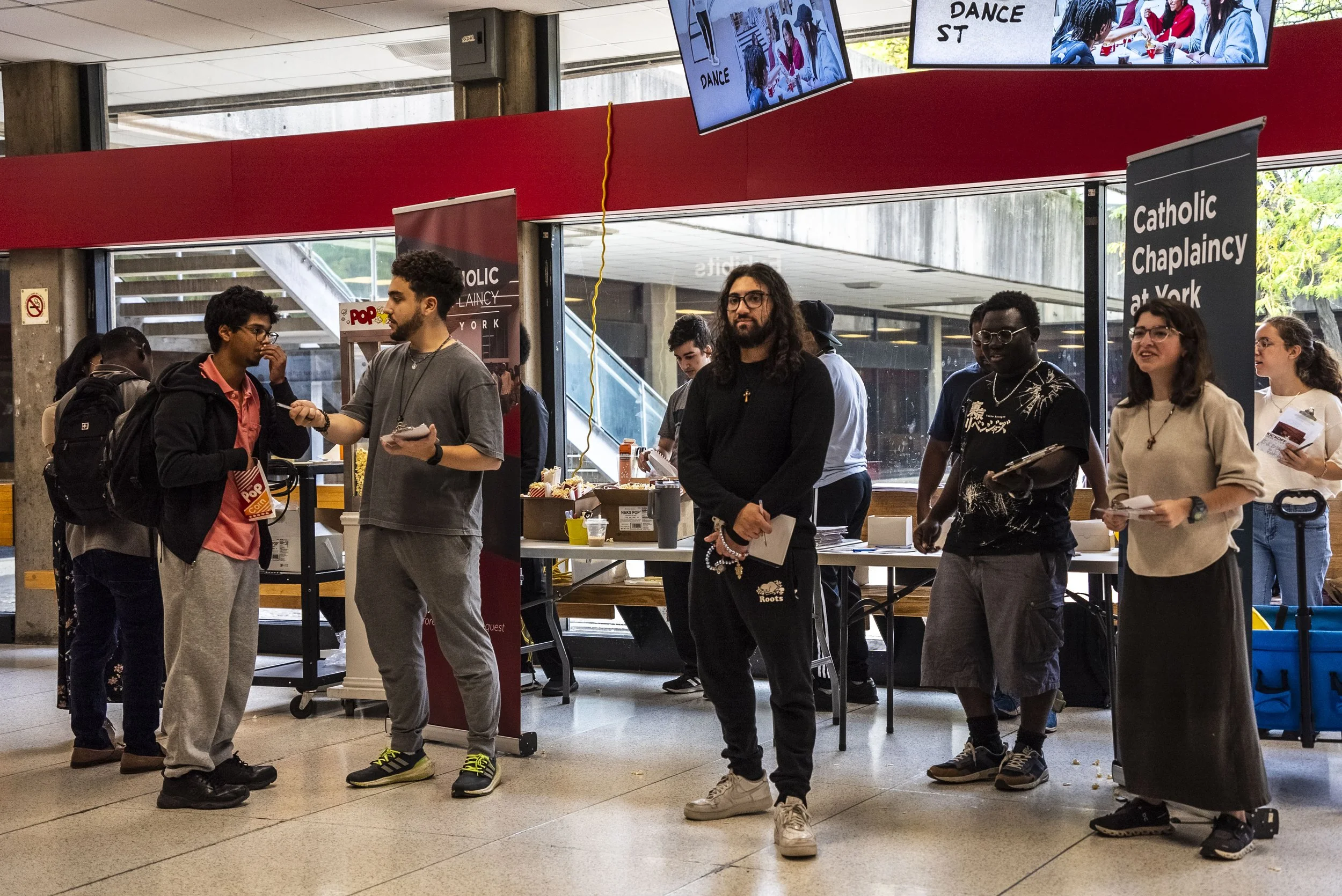 A group of diverse people waiting in line at a ticket counter inside a building, with digital screens displaying images of people dancing, and signs indicating it is for a Catholic chaplaincy at York.