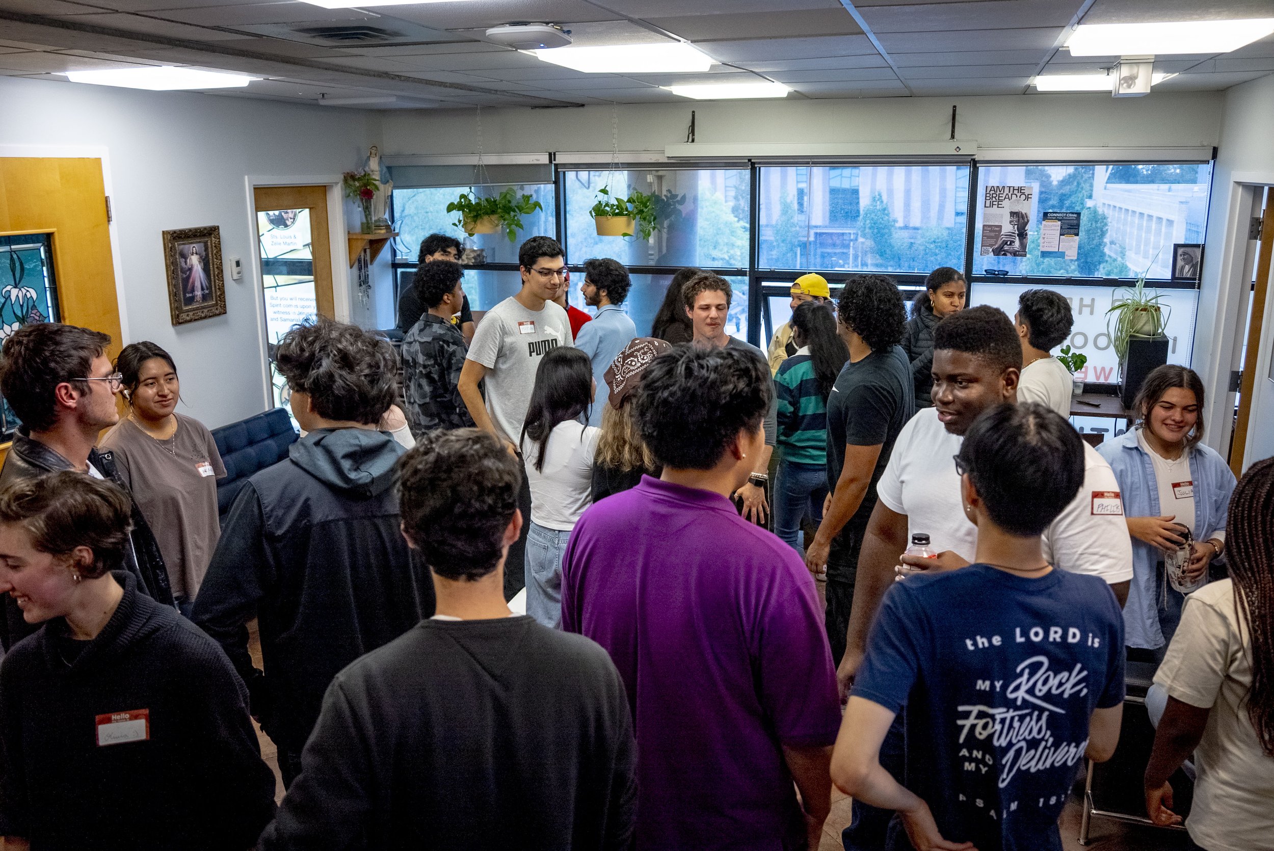A group of diverse young adults gather indoors, some engaging in conversation while others are smiling or listening. The room has large windows, hanging plants, and various posters and artwork on the walls.