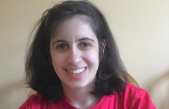 A smiling woman with curly dark hair, wearing a pink top, in front of a light-colored background.