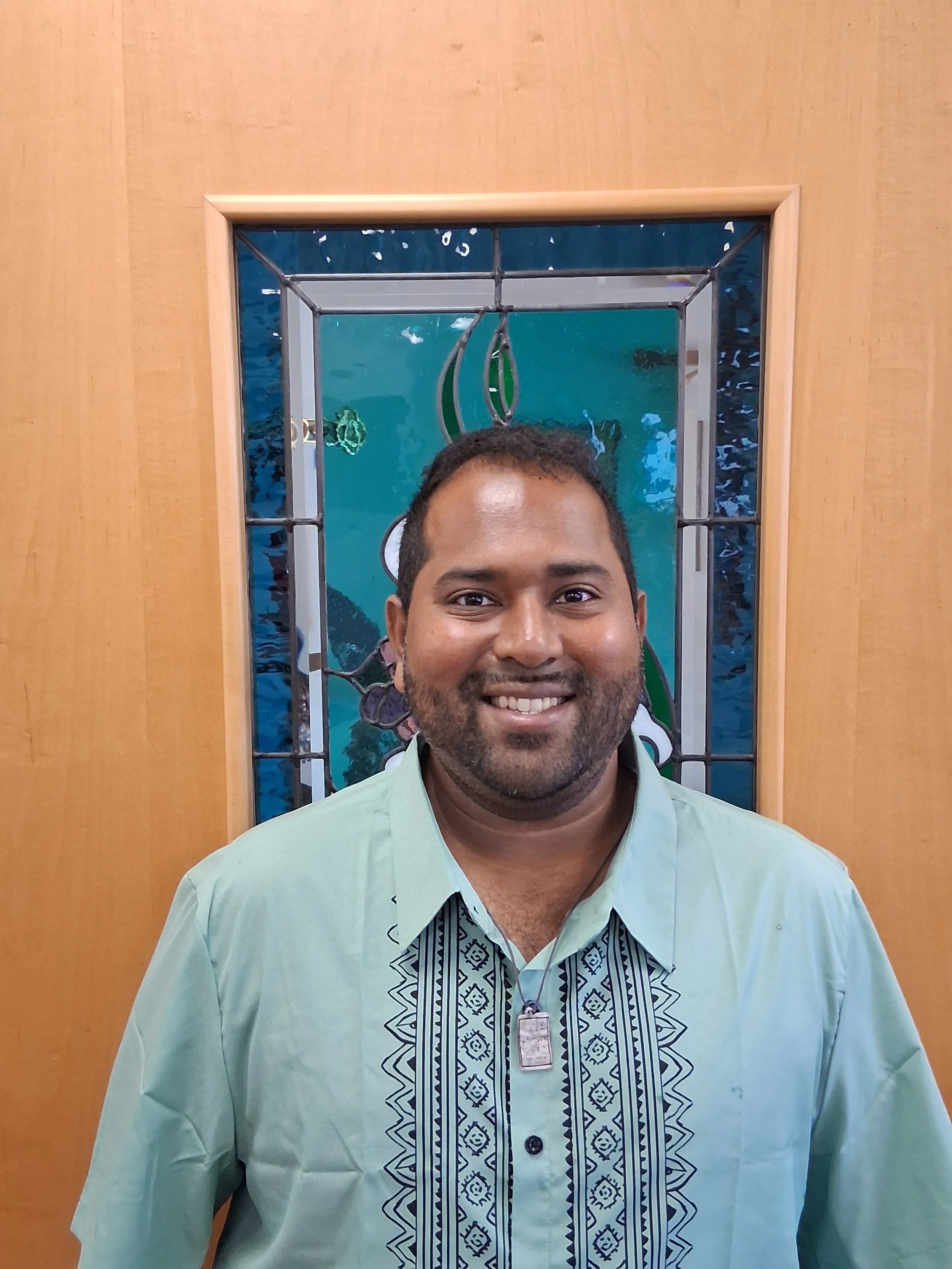 A man wearing a light green shirt with embroidered dark blue and white patterns in the front, smiling, standing in front of a wooden door with a mirror behind him. The mirror reflects a blue wall with some fish and a part of a fish tank.