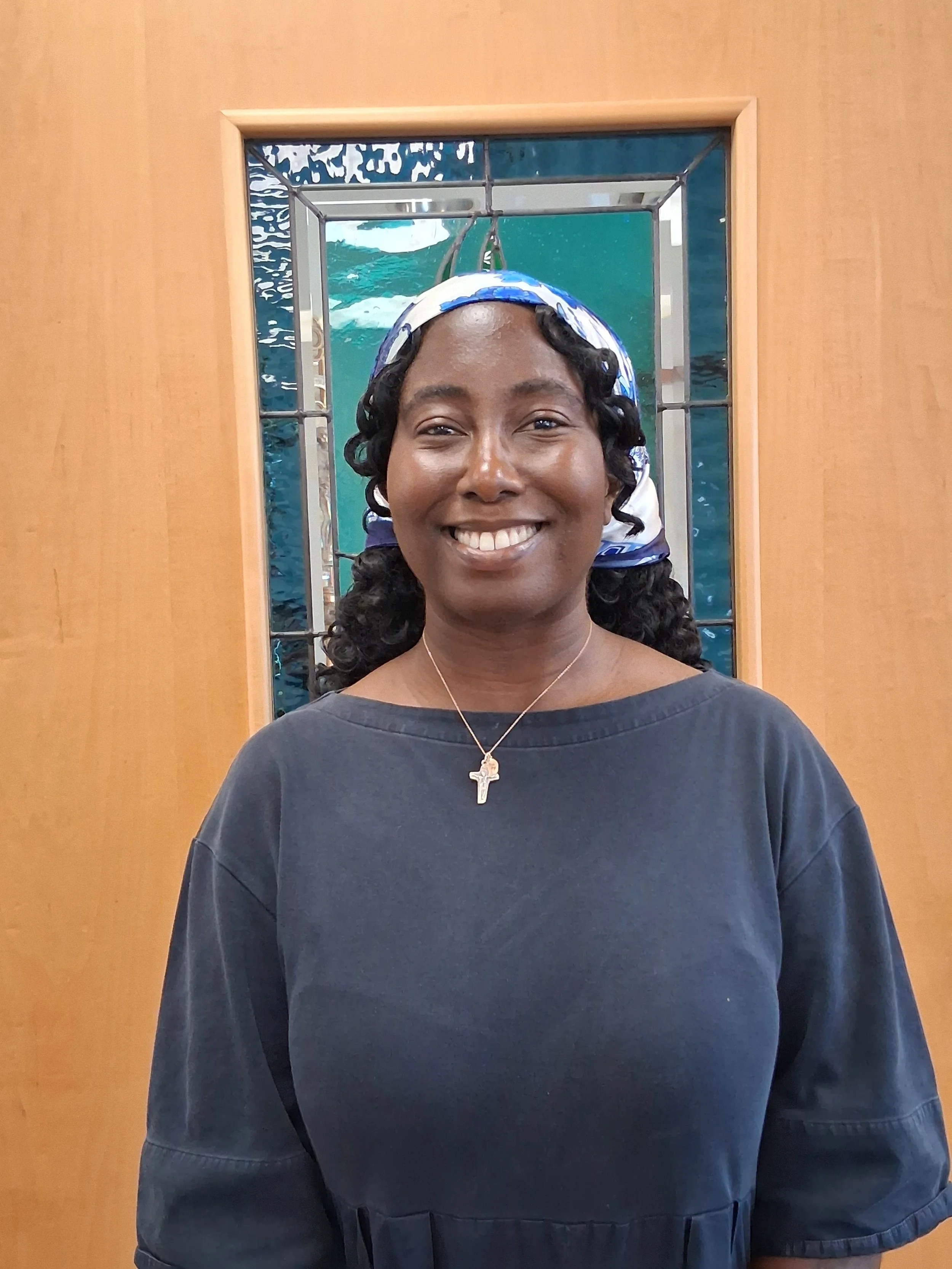A woman smiling in front of a mirror with a window behind her, carved wood wall, wearing a dark shirt, a necklace with a cross, and a colorful headscarf.