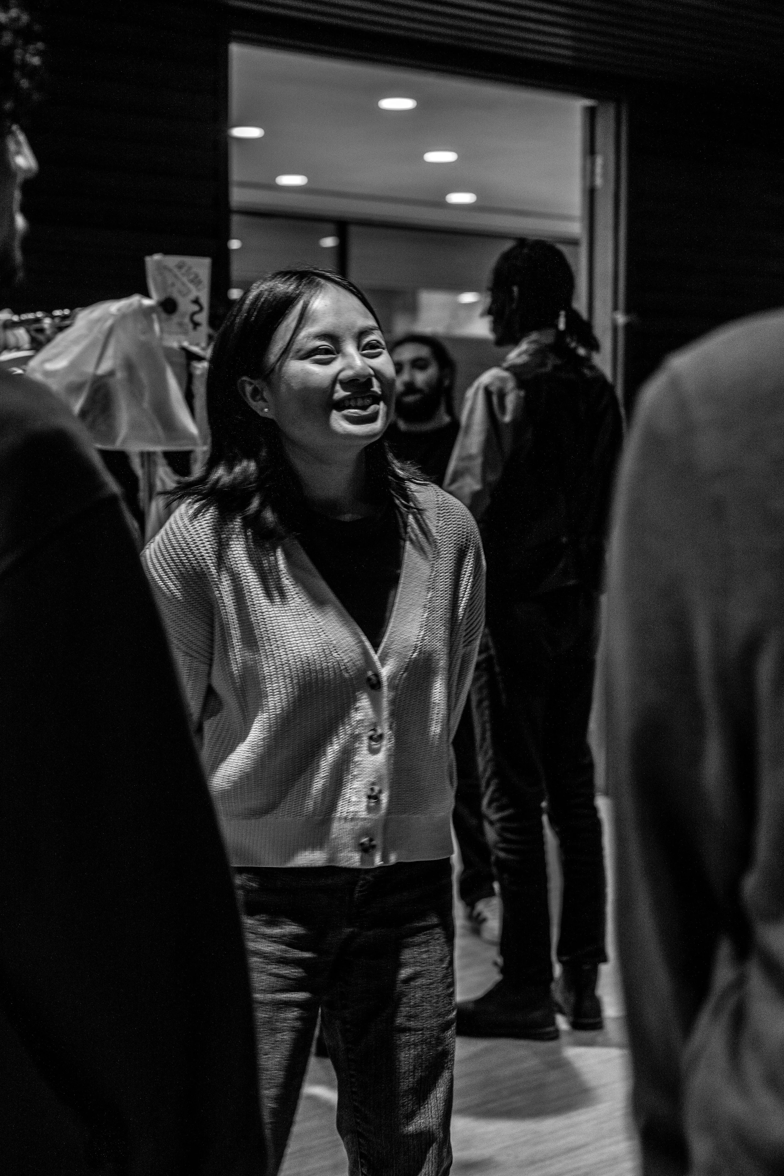 Black and white photo of a woman smiling and talking to someone in a social setting. Several other people are visible in the background, some engaged in their own conversations.