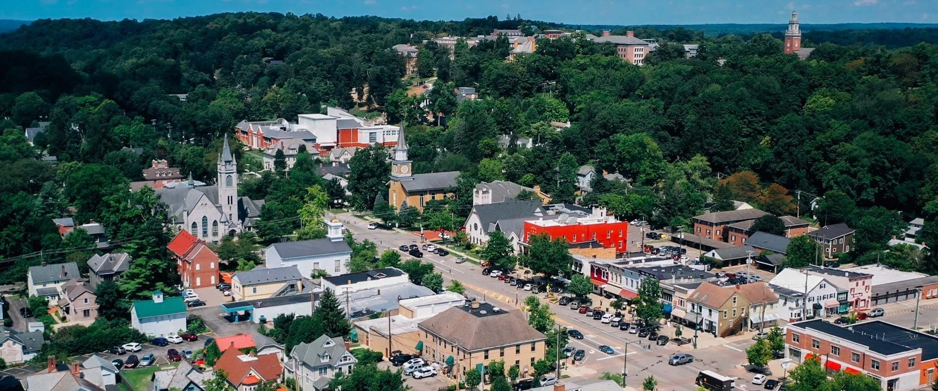 Aerial view of a small town surrounded by green trees and hills, featuring churches, colorful buildings, and parking lots.
