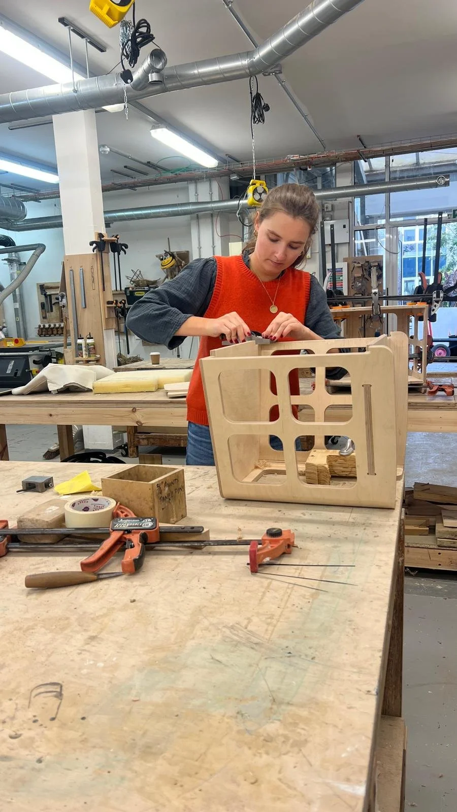 A woman working on a woodworking project at a workshop, assembling or sanding a wooden piece with various tools and materials scattered on the table.