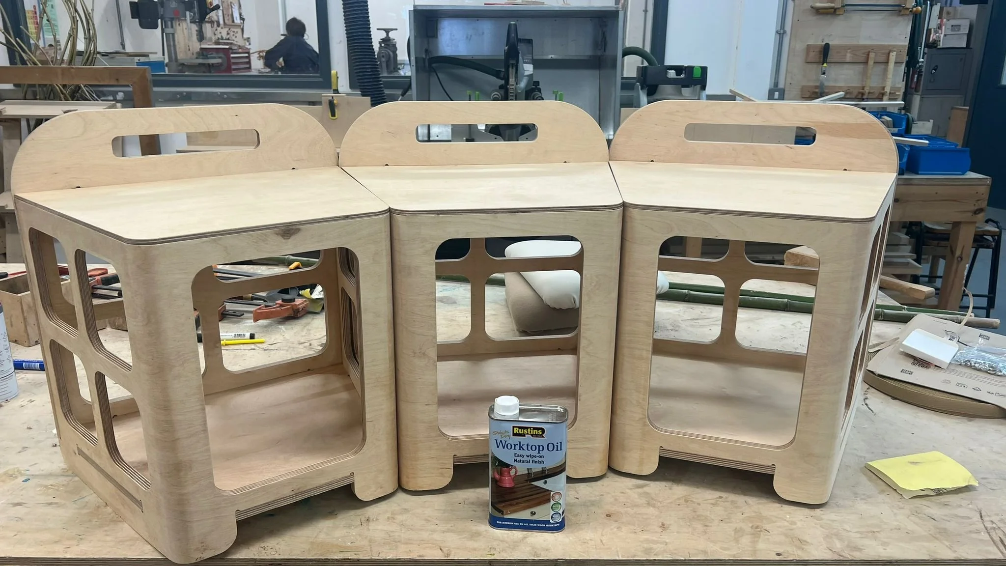 Unfinished wooden furniture, possibly a set of shelves or a storage unit, on a workbench in a woodworking shop, with tools and equipment in the background.