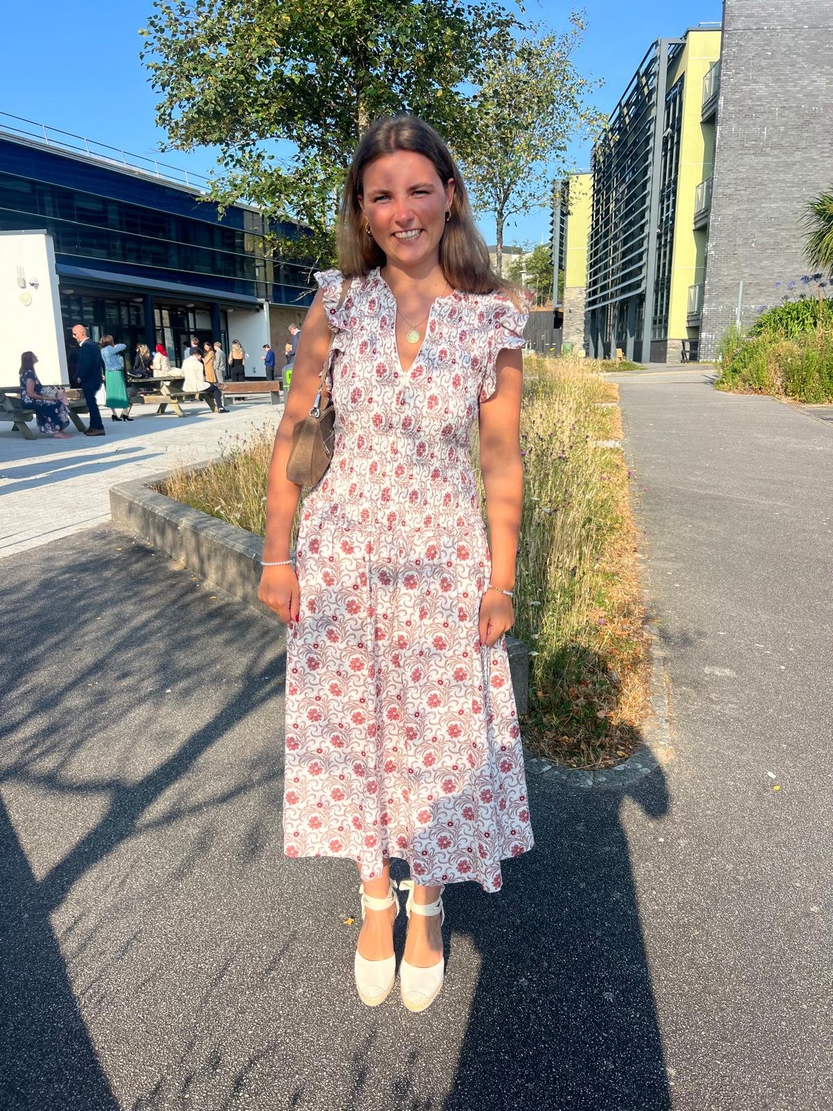 A woman stands outdoors on a sunny day, smiling and wearing a long, patterned dress with white high heels, carrying a small purse, with a modern building and a group of people in the background.