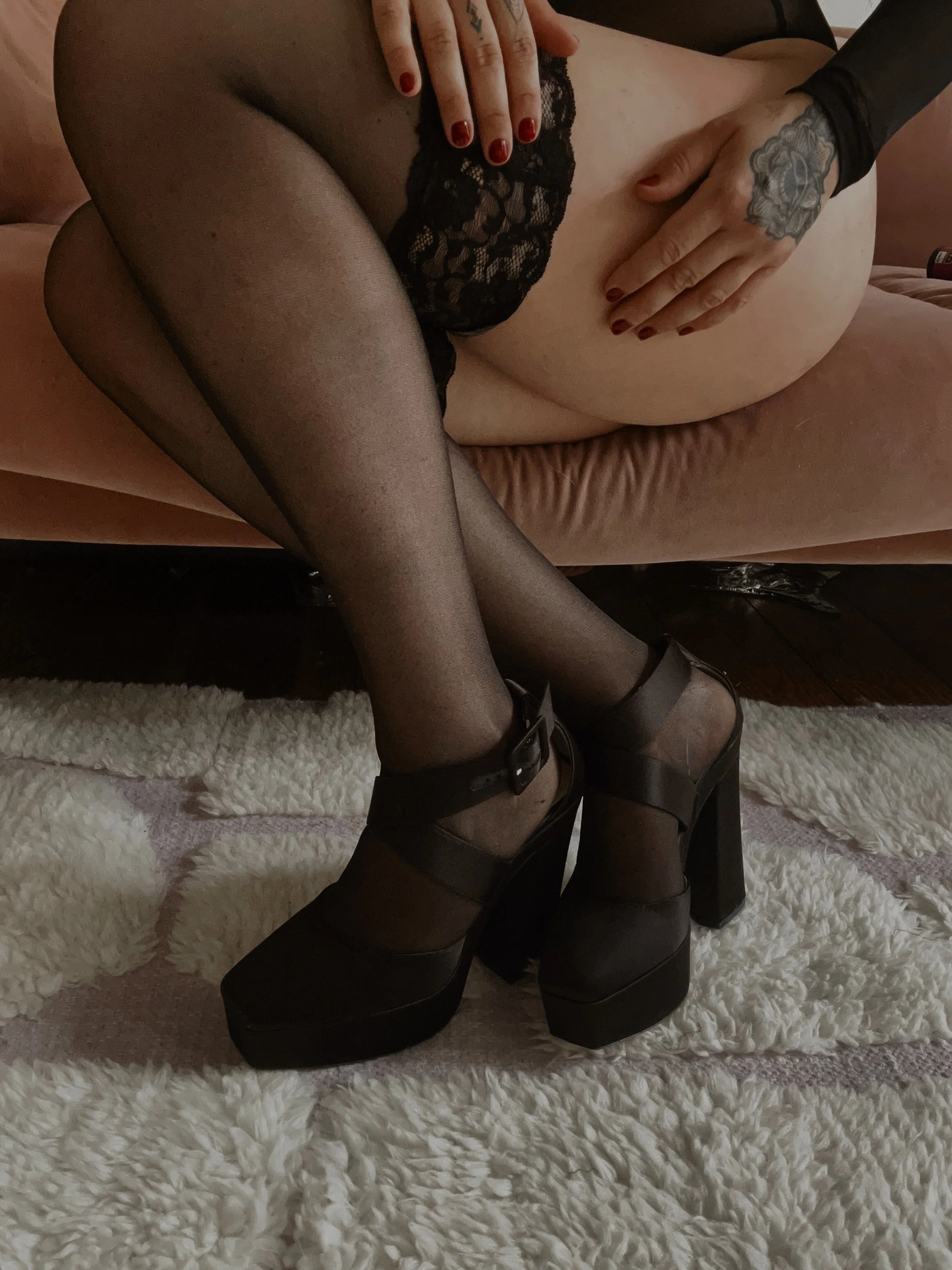 Close-up of a person sitting on a soft rug, wearing black sheer tights, high platform black shoes, and black lace underwear. The person has tattoos on their hands and a tattoo on their wrist, with red nail polish on their fingernails. The scene is indoors with a pinkish background.