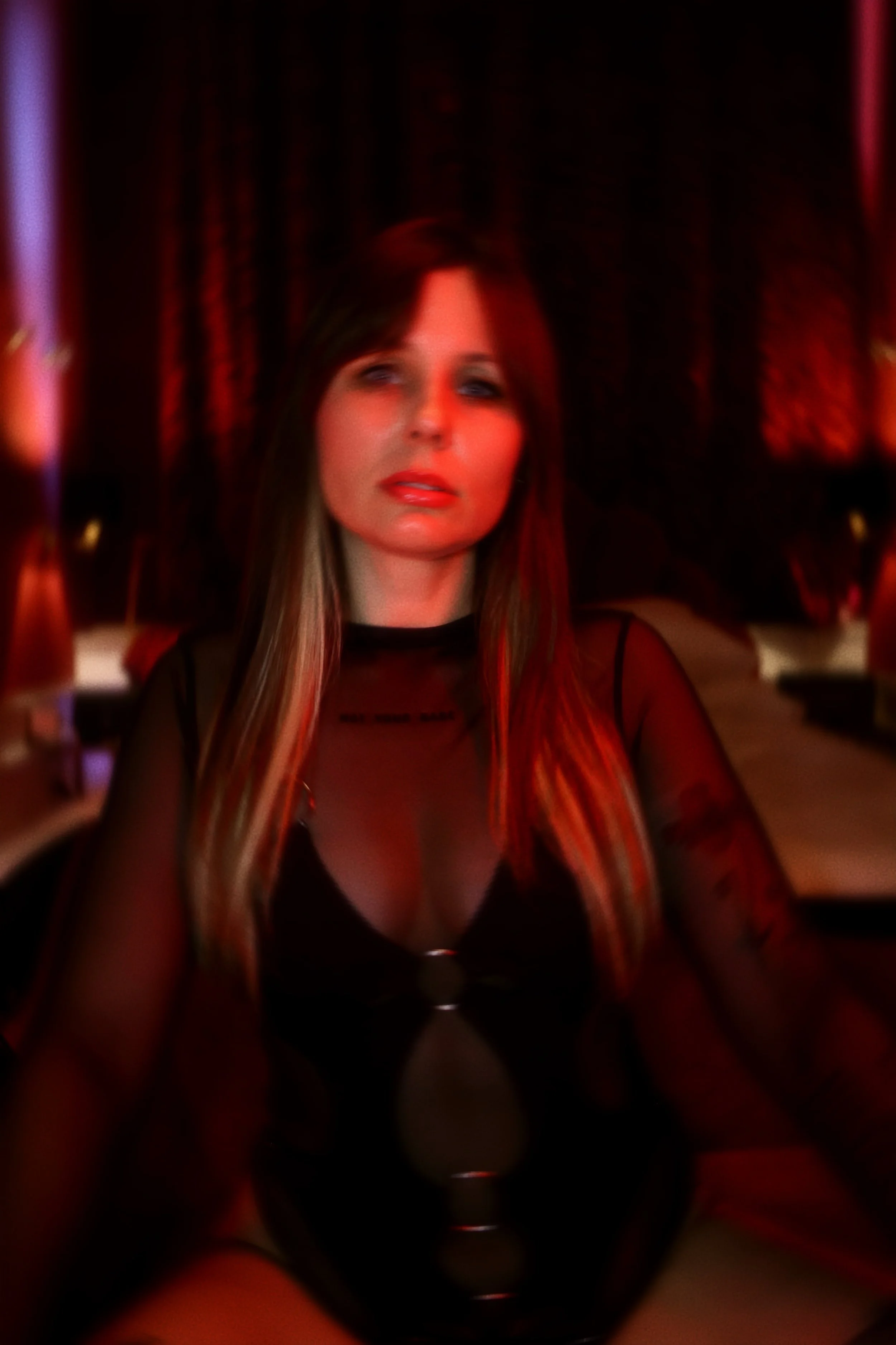 A woman with long hair and makeup sitting at a table in a dimly lit room with red and purple lighting.