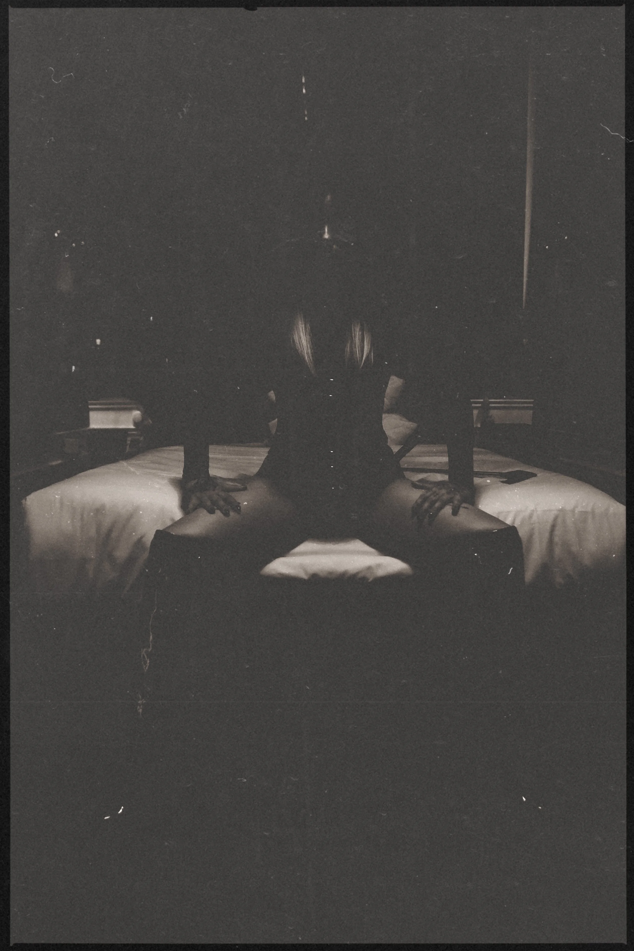 A person with long hair is lying on a bed, positioned with their head near the edge, facing downward, with their back arched and legs spread. The photo is in black and white.