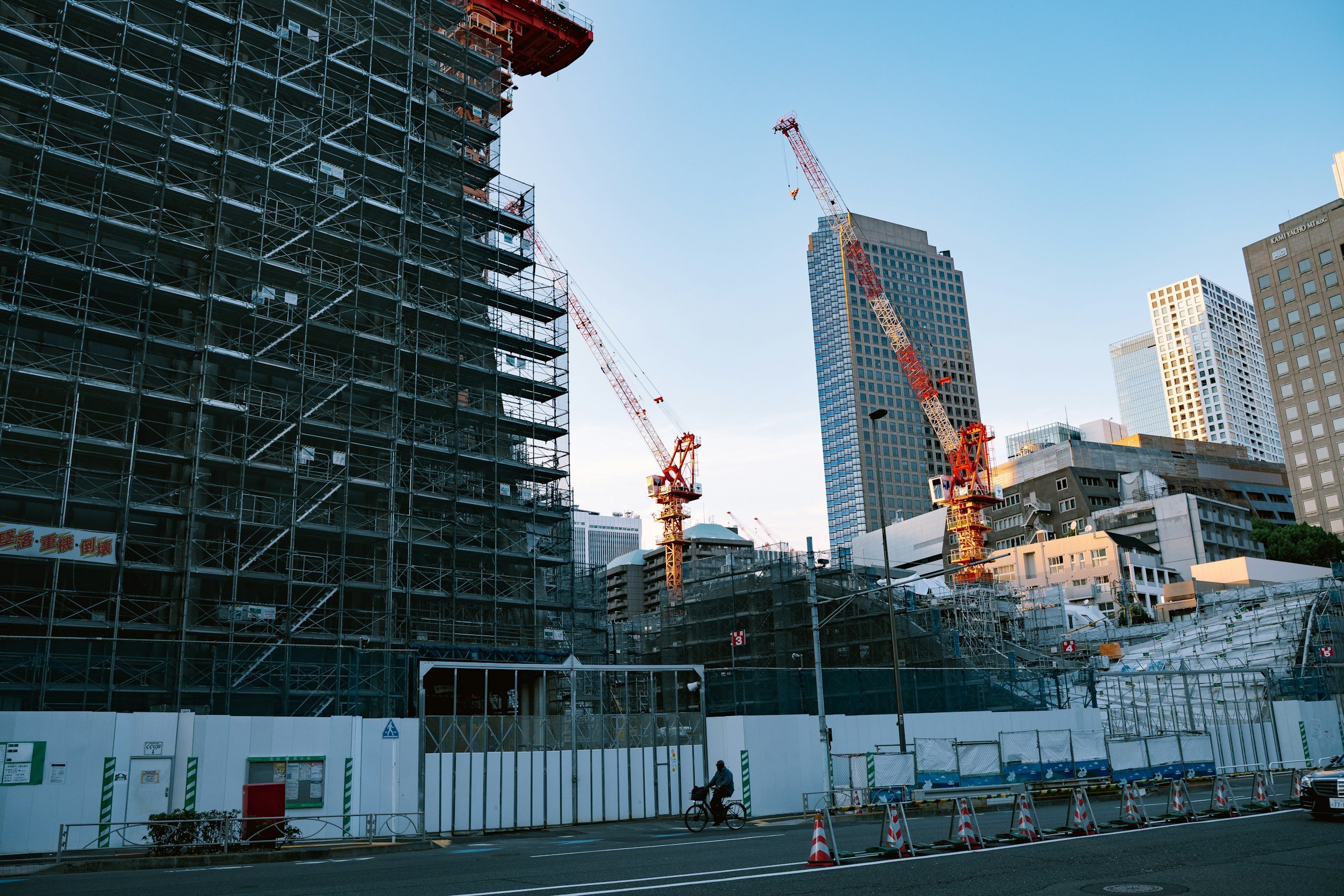 Construction site in an urban area with scaffolding around a building under construction, several construction cranes in use, and high-rise buildings in the background. A person riding a bicycle and another walking are visible on the street in front of the construction zone, which is enclosed by a white fence.