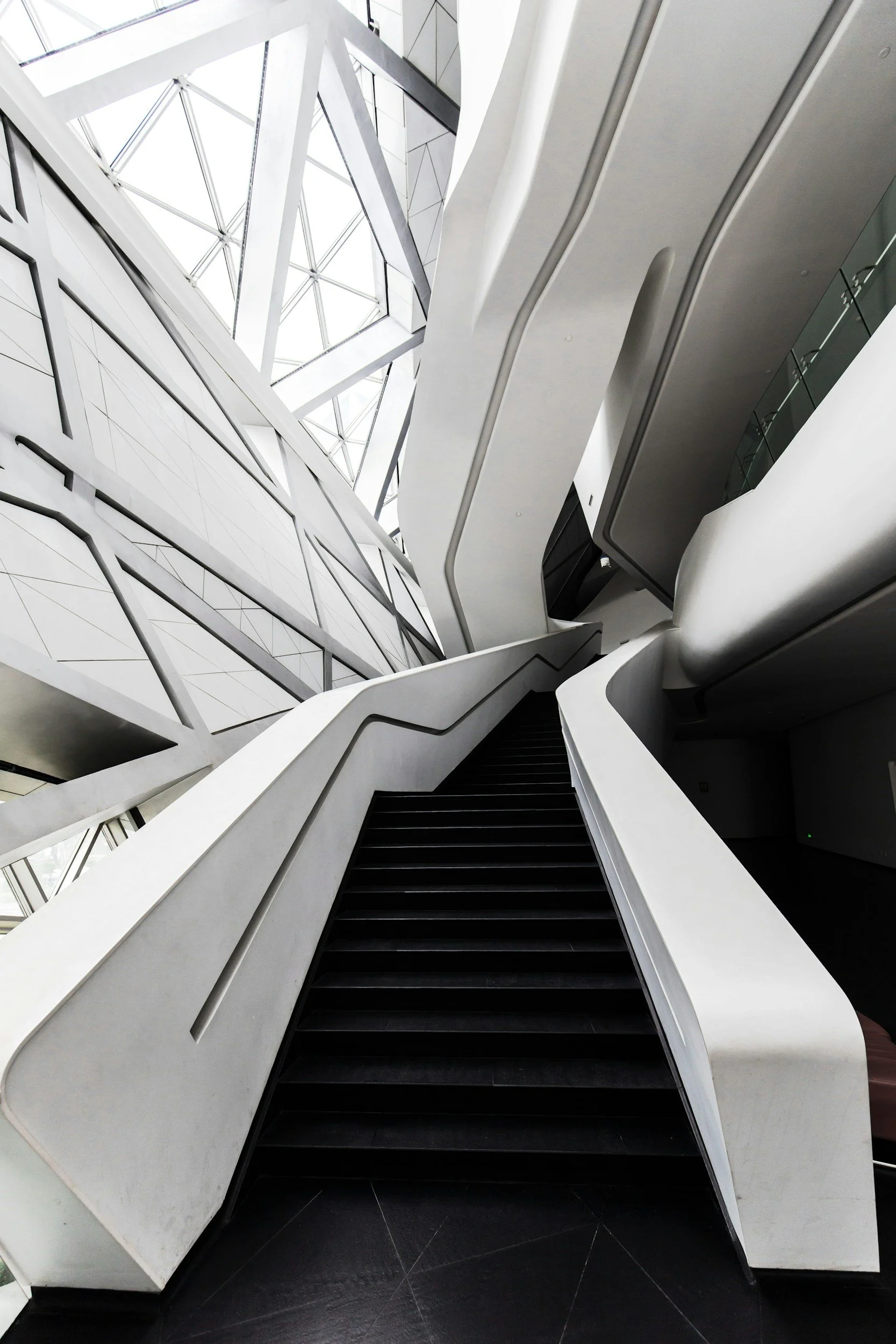 Modern architectural interior featuring a black staircase with white railings, curved white walls, and a geometric glass ceiling allowing natural light to illuminate the space.