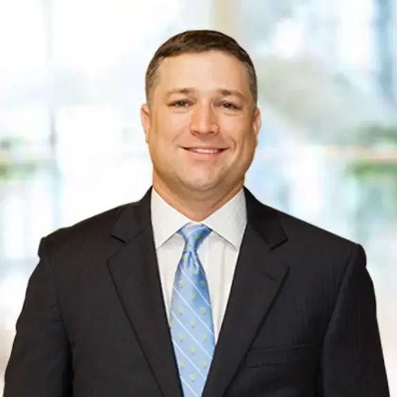 Chris Epps, founder of Clear Claim Consulting, in a black suit, white shirt, and light blue tie, standing indoors with a blurred window background.