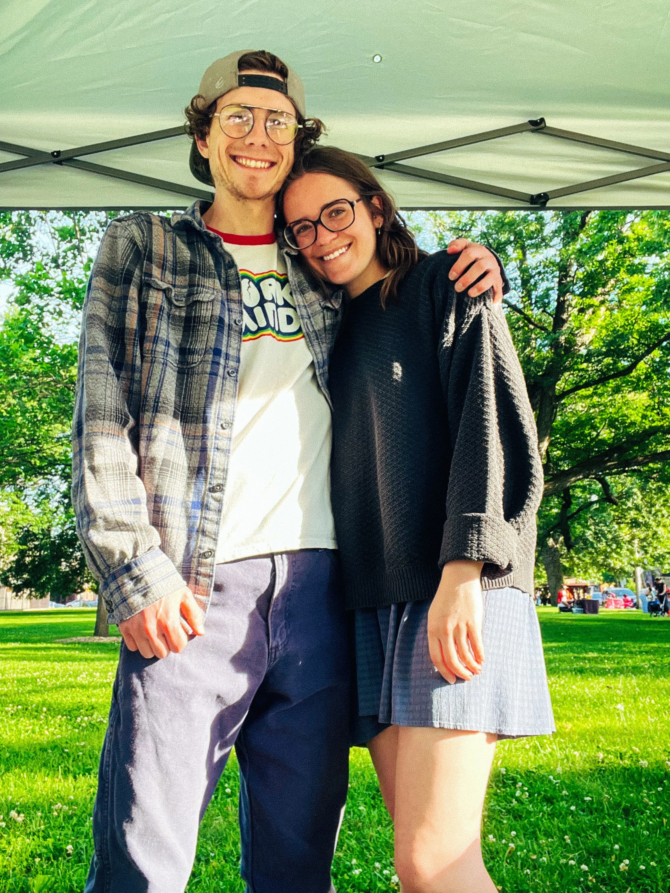A young man and woman smiling and hugging in a park under a white canopy, with green grass and trees in the background.
