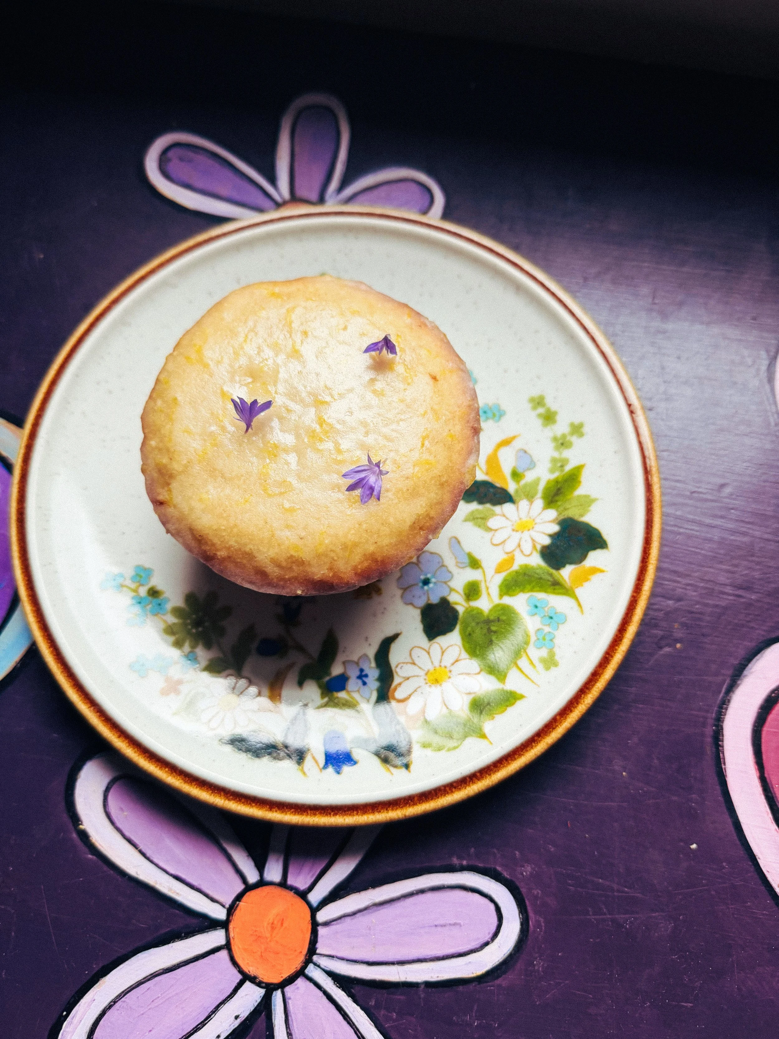 A bowl with a floral pattern and gold rim containing a muffin decorated with purple edible flowers, placed on a purple placemat with a flower design.