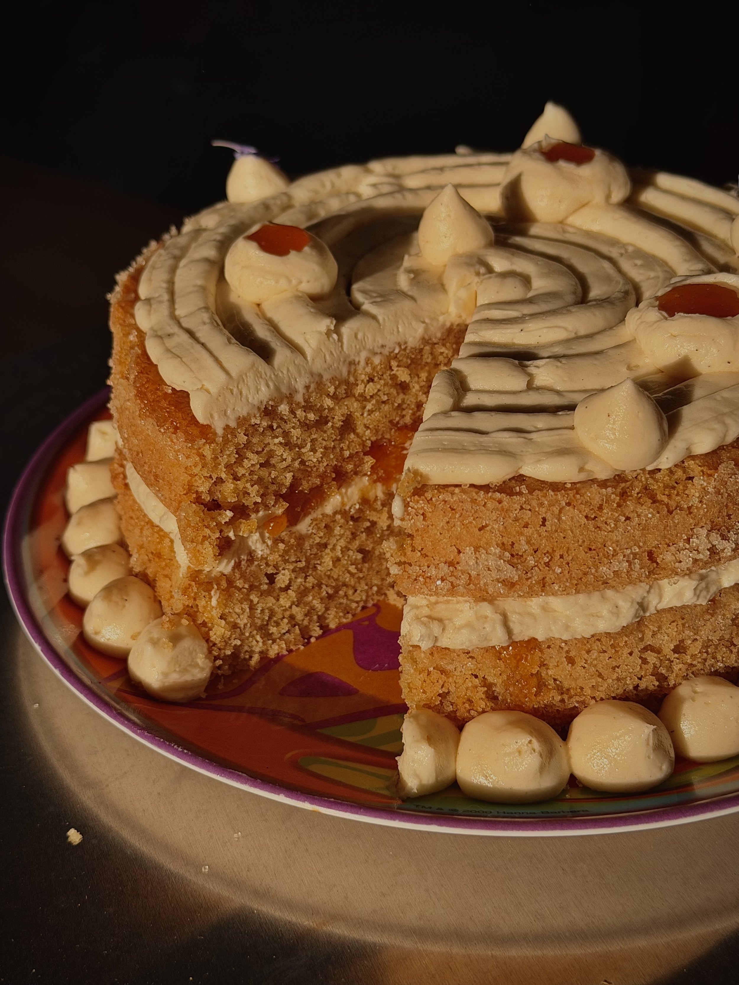 A slice of cake with white frosting decorated with white chocolate chips and red jelly on top, on a colorful plate with more white chocolate chips around it.