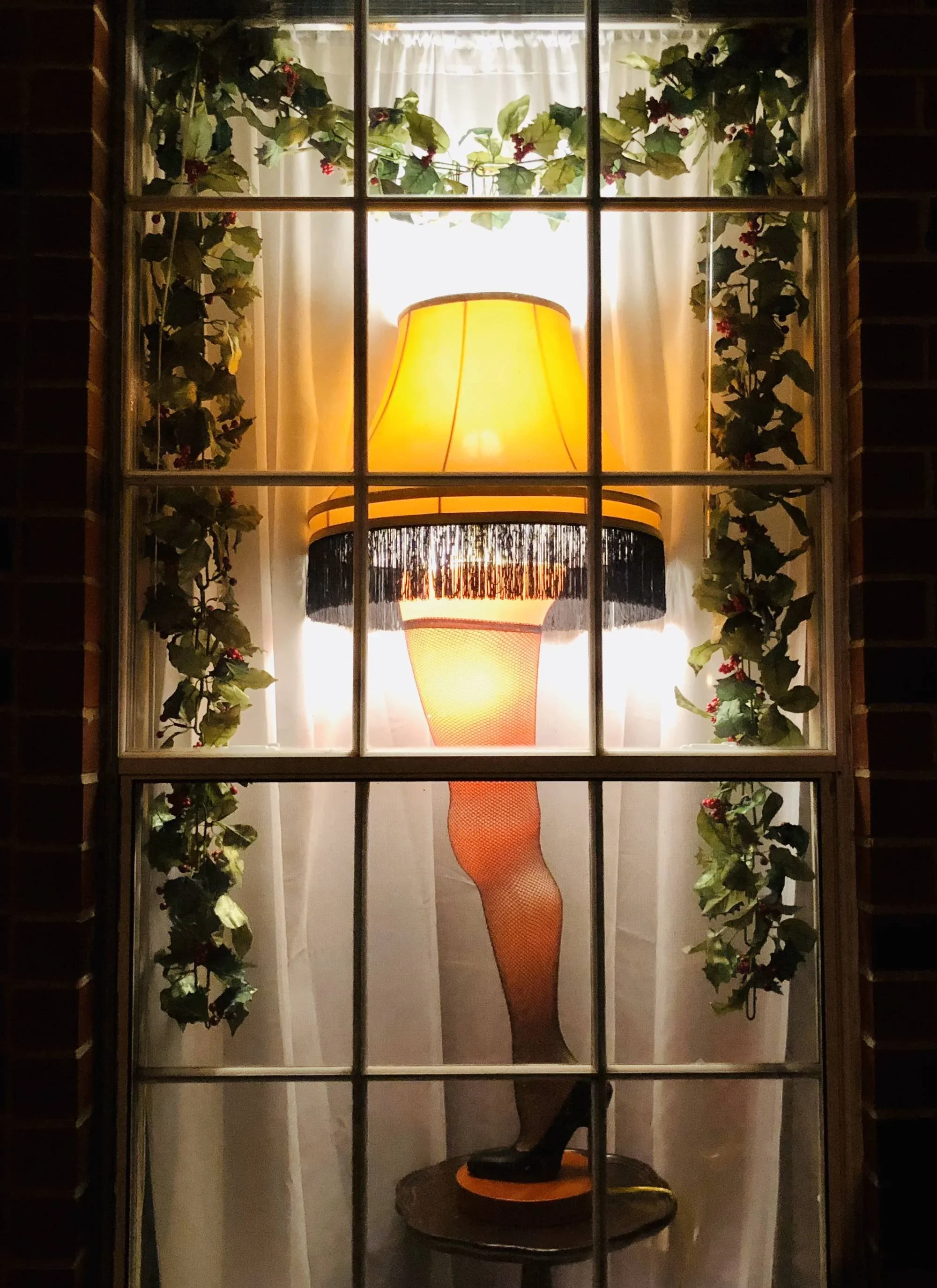 A window with a grid, showing a side view of a large, orange, textured lampshade with black fringe and a black base, sitting on a small black table. There are white curtains behind the lamp, and the window is decorated with green ivy with red berries.