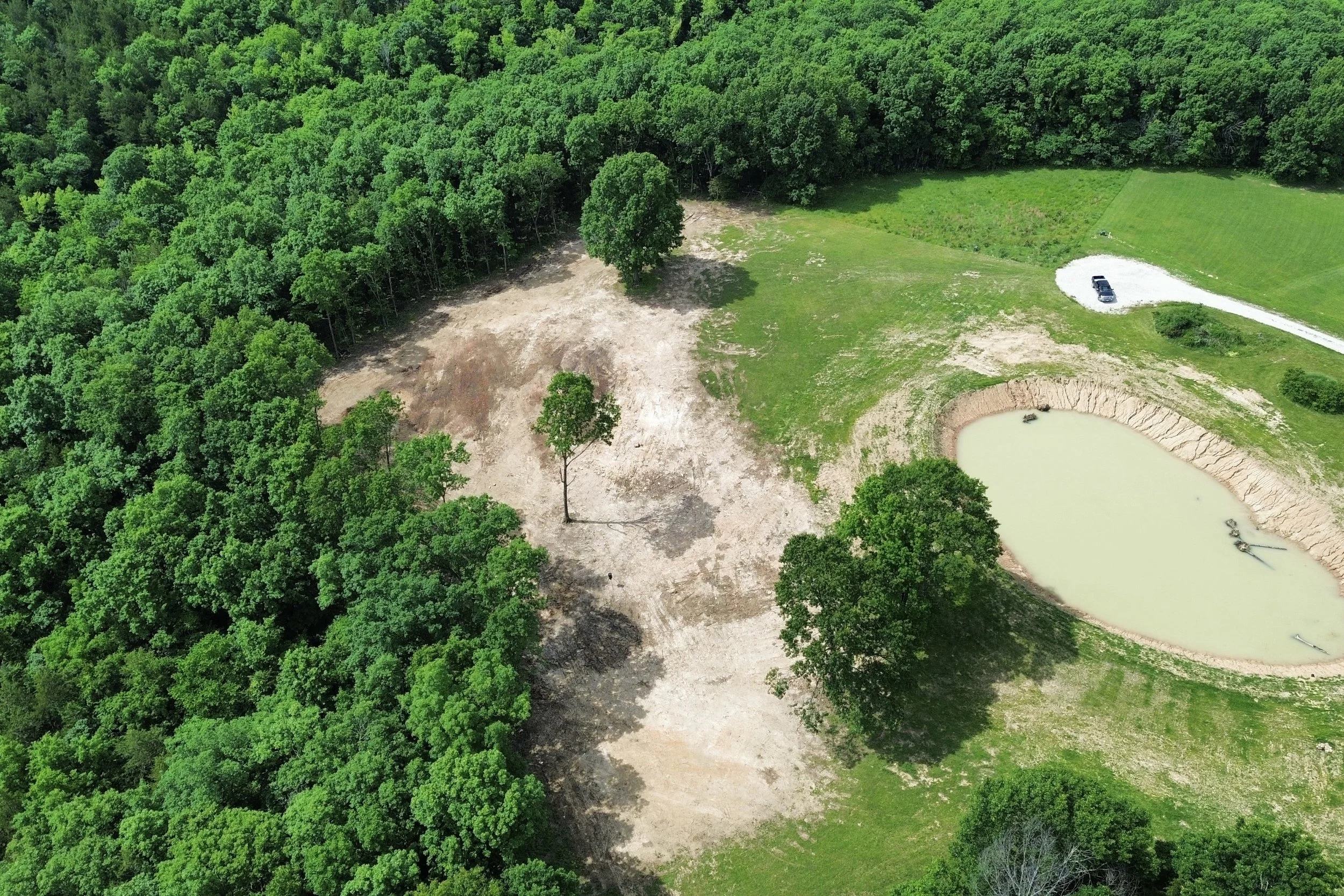 Aerial view of a large area partly cleared of trees with green forest surrounding it, a small pond, and a gravel driveway with a car parked on it.