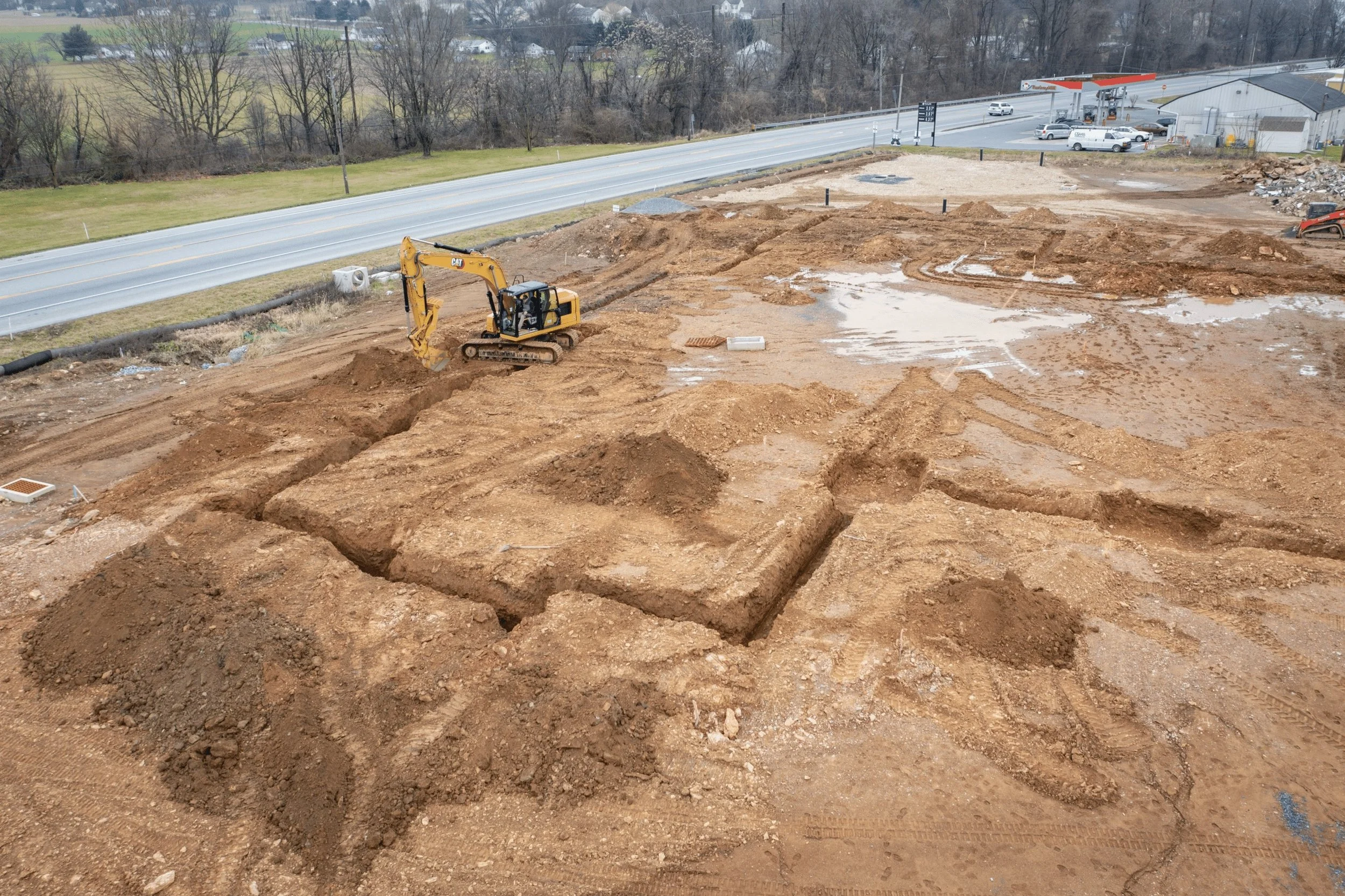 Construction site with excavator digging earth, with a commercial area, gas station, and roads in the background.