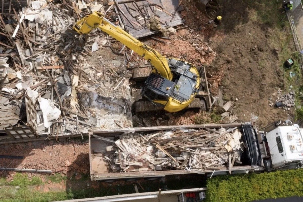 An aerial view of a construction site with a yellow excavator demolishing a building, debris scattered around, and a truck transferring debris.