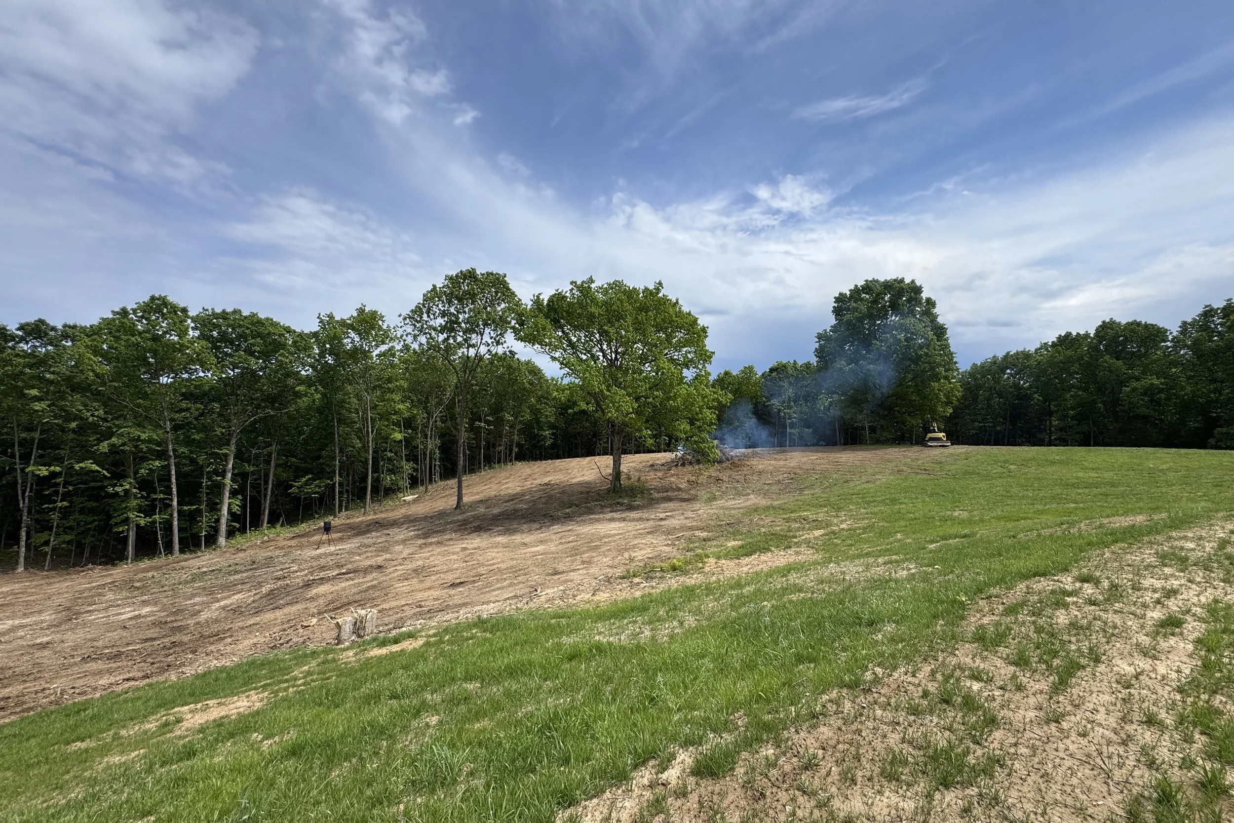 A landscape with a grassy foreground, a partially cleared wooded area, and a blue sky with clouds. A tractor is visible on the right side, and there is smoke or dust rising from the cleared area.