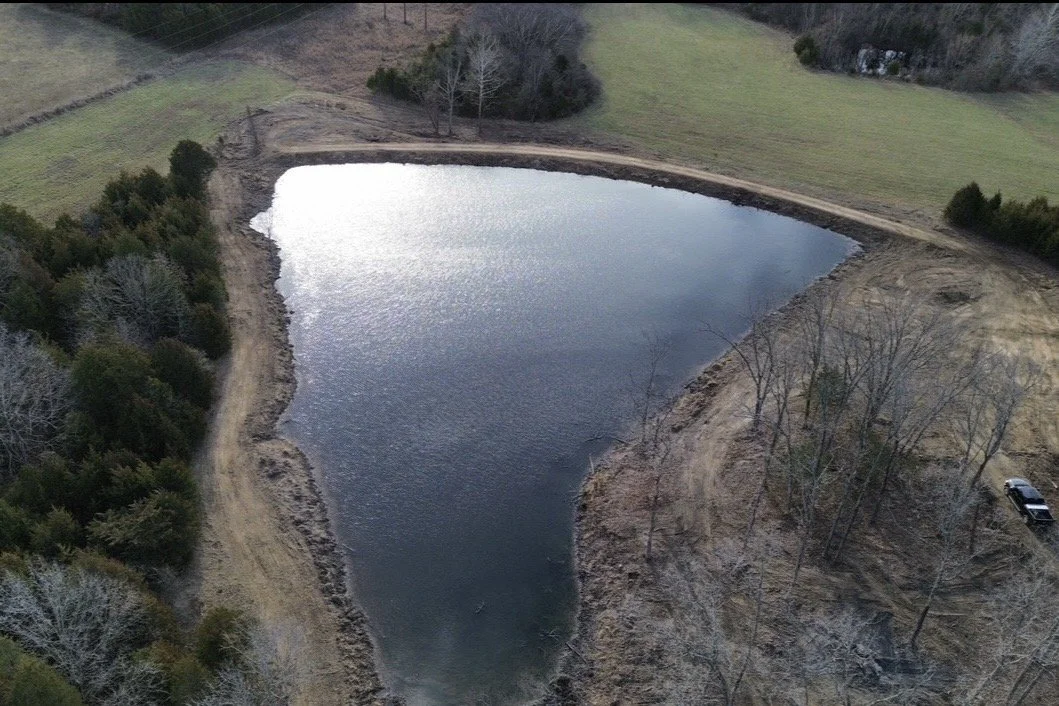 A small pond surrounded by dirt and sparse trees in a rural landscape