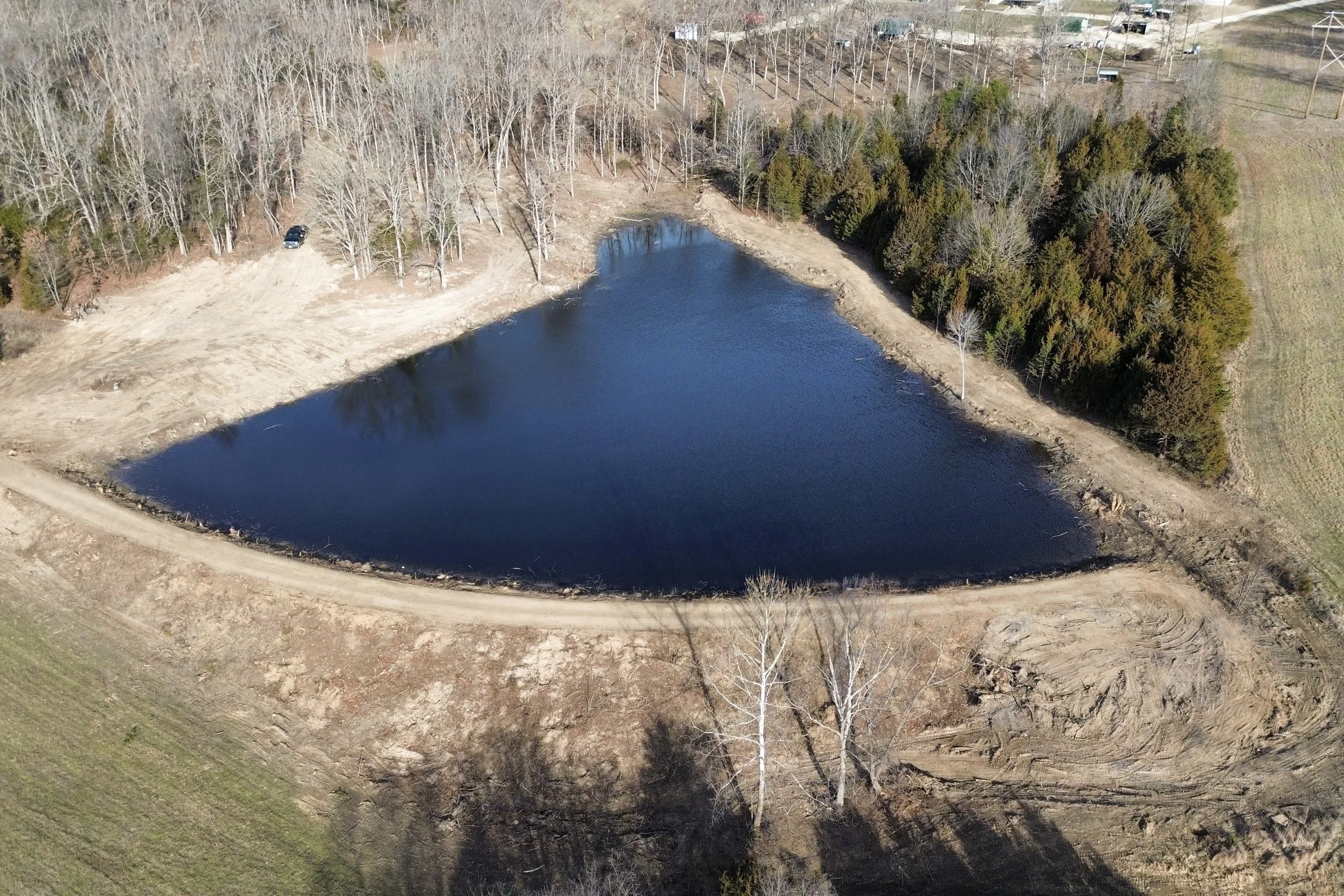 An aerial view of a small lake surrounded by a dirt path and trees, with a few buildings and power lines in the background.