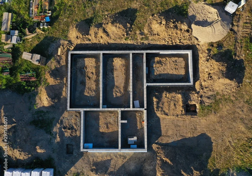 Aerial view of a house foundation under construction with concrete walls forming the outline of the building's layout, surrounded by dirt and construction materials.