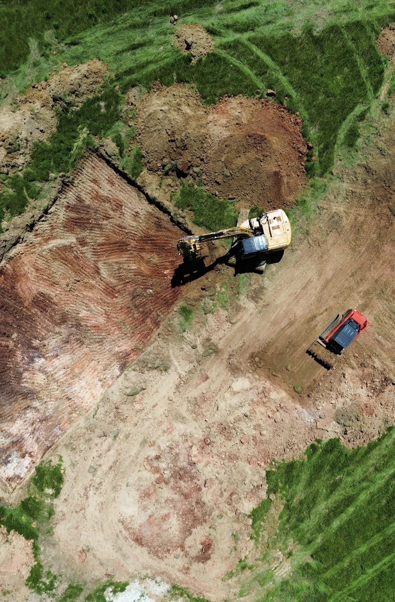 An aerial view of construction equipment and excavated soil in a grassy area with trees.