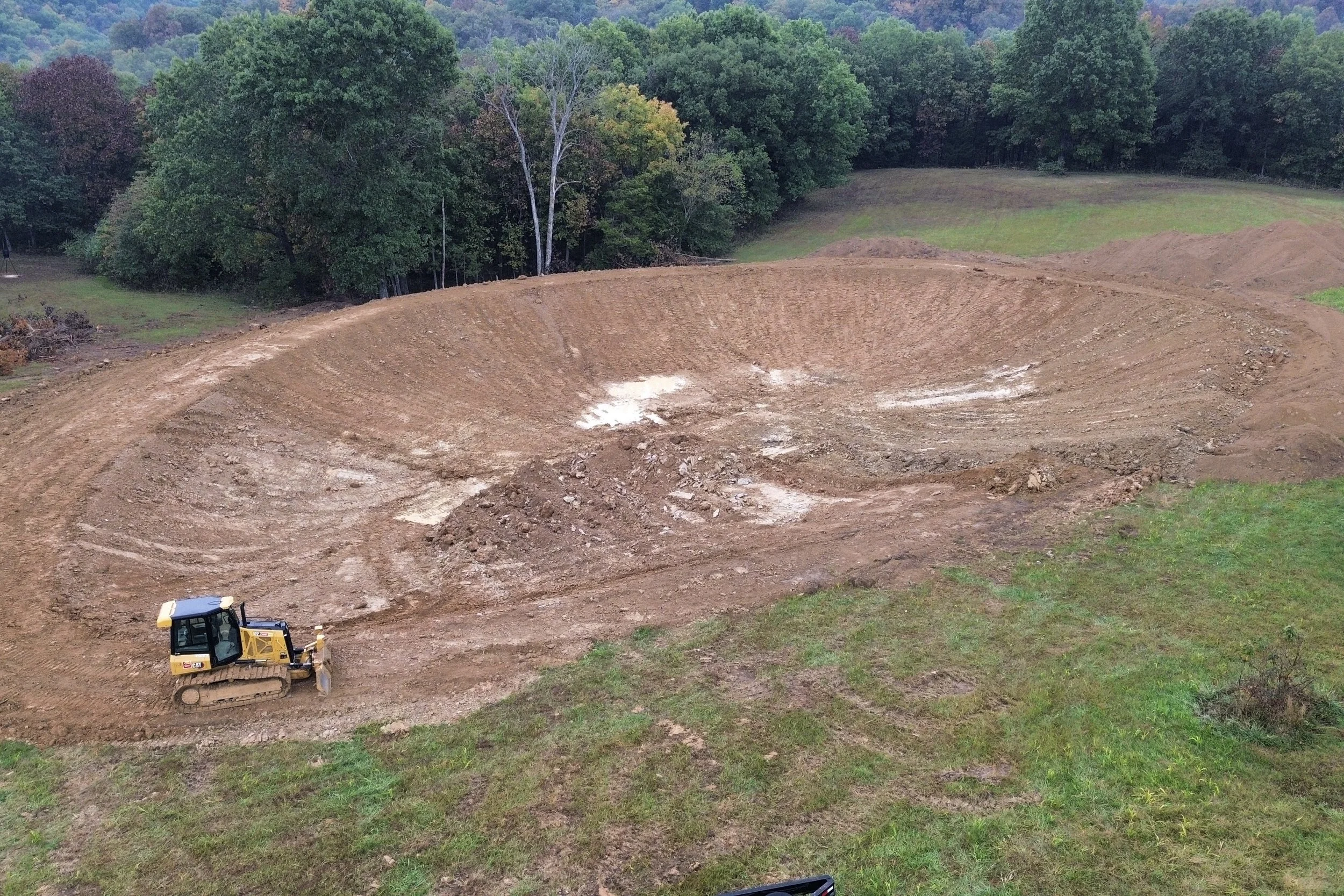 A yellow bulldozer working on a construction site in a grassy area near a forested hill.