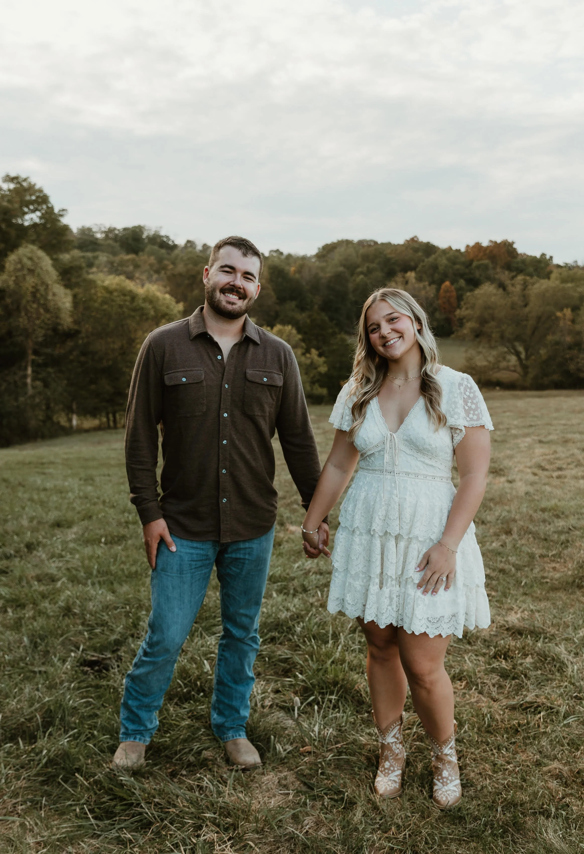 A smiling man and woman holding hands outdoors in a field, with trees and a cloudy sky in the background.