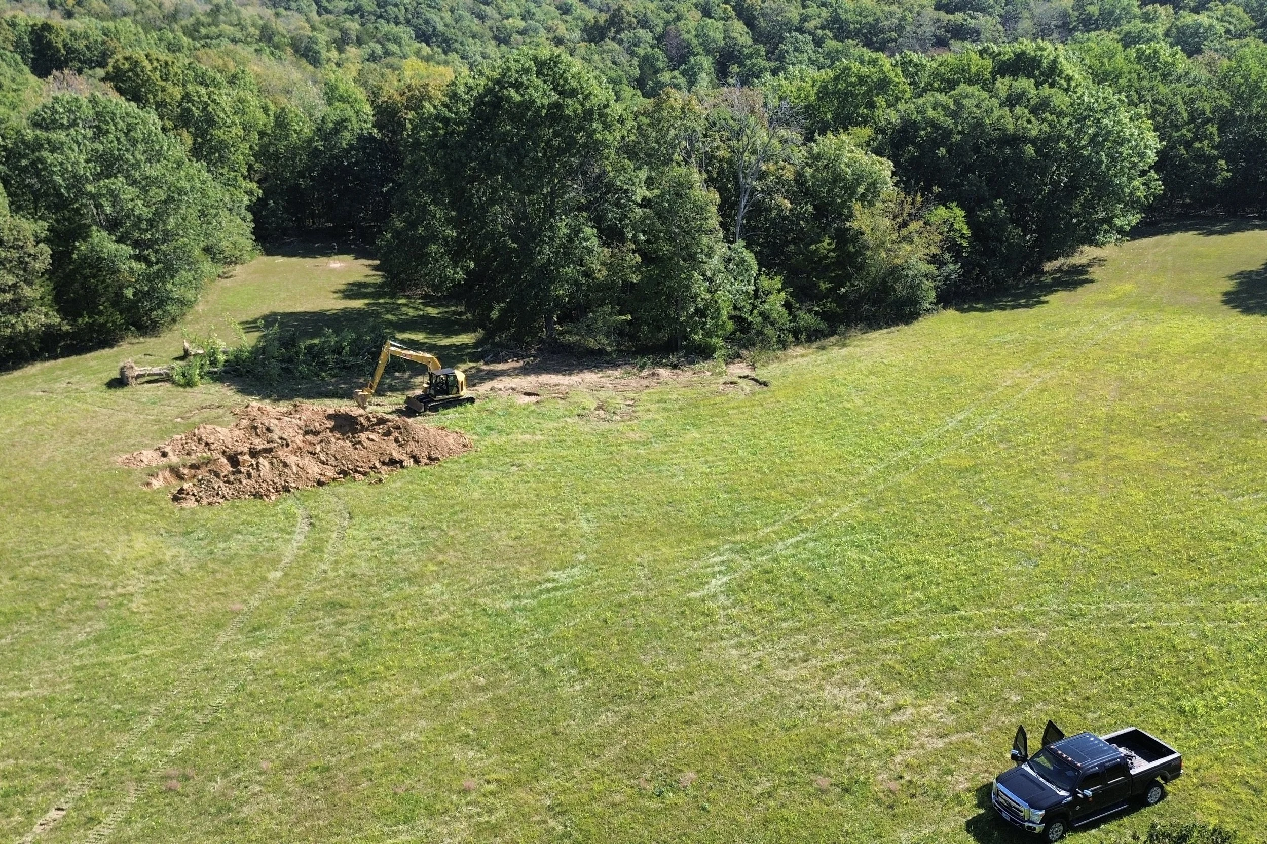 A construction site in a grassy field with a yellow excavator digging a hole near a pile of dirt, surrounded by trees in a lush green landscape.