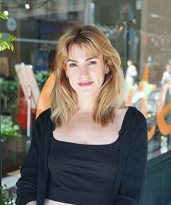 Une femme aux cheveux blonds et aux yeux clairs, portant un haut noir, se tenant devant une vitrine de magasin avec des objets colorés à l'intérieur.
