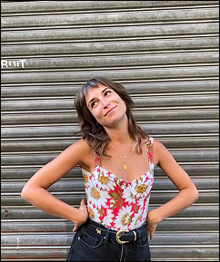 Jeune femme posant devant un rideau en métal, souriante, portant un débardeur floral et un jean noir.