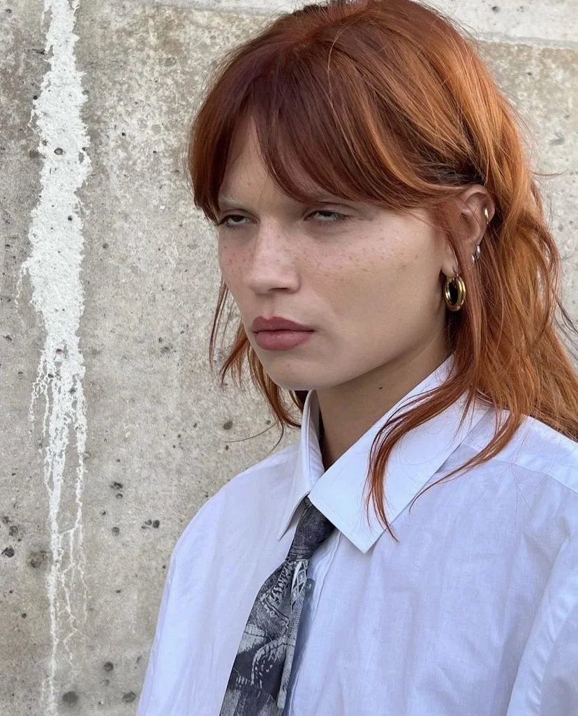 Jeune femme avec des cheveux roux, portant une chemise blanche et une cravate noire, posant devant un mur en béton.