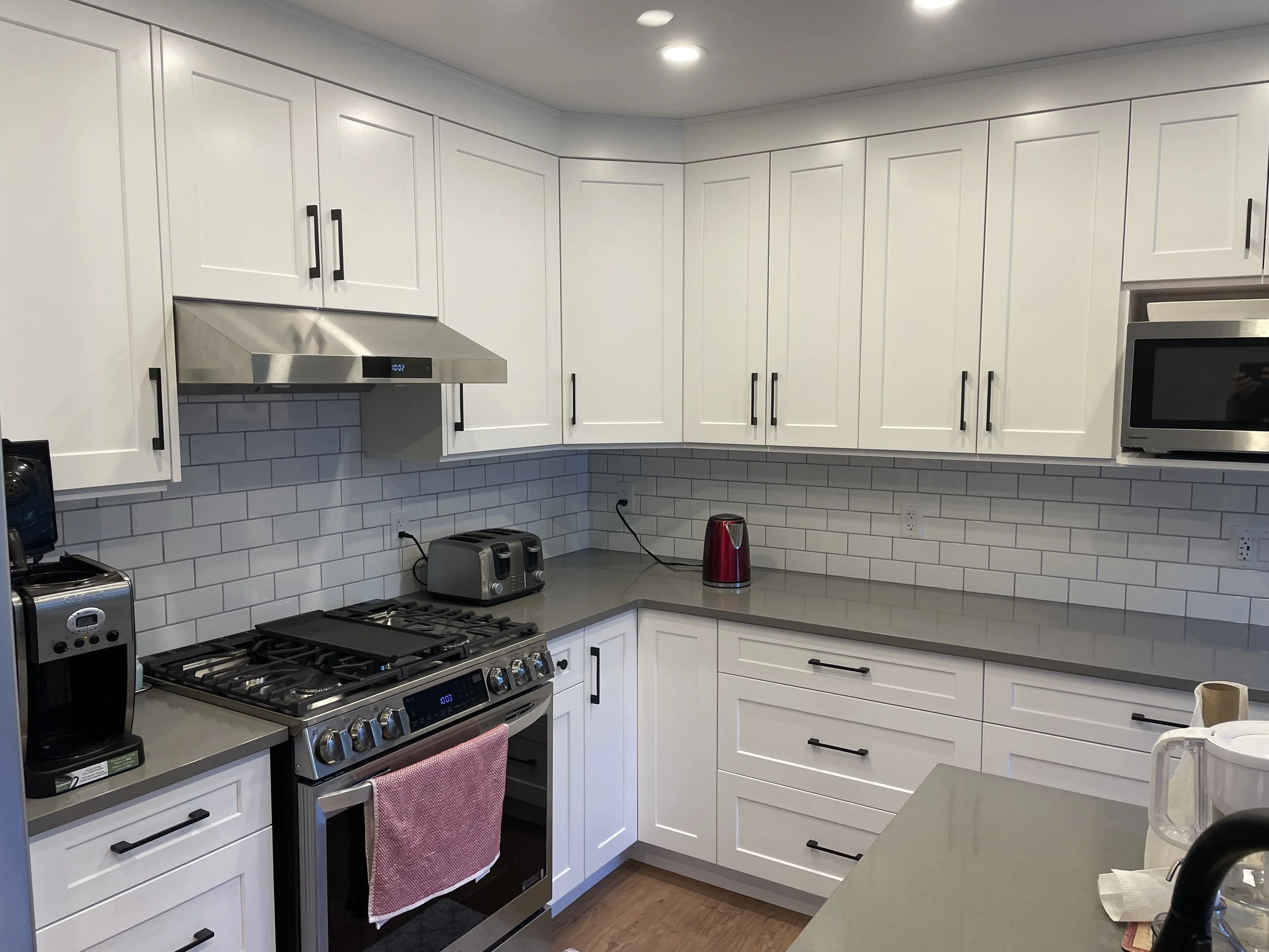 A modern kitchen with white cabinets, a stainless steel stove and range hood, a microwave, and various small appliances on the countertop.