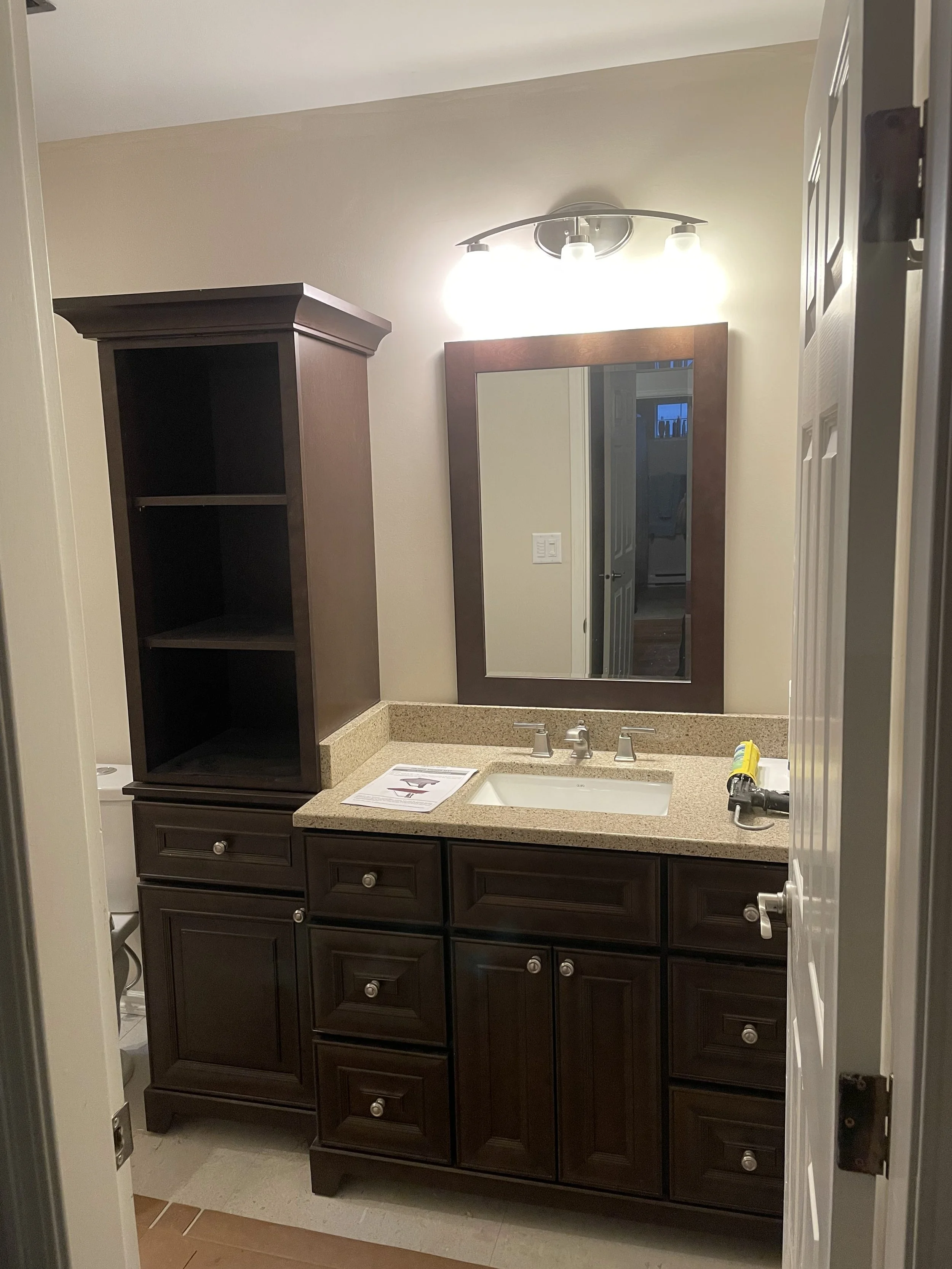 Bathroom with dark wood vanity, beige countertop with a sink, large mirror with wooden frame, and above light fixture with three bulbs.