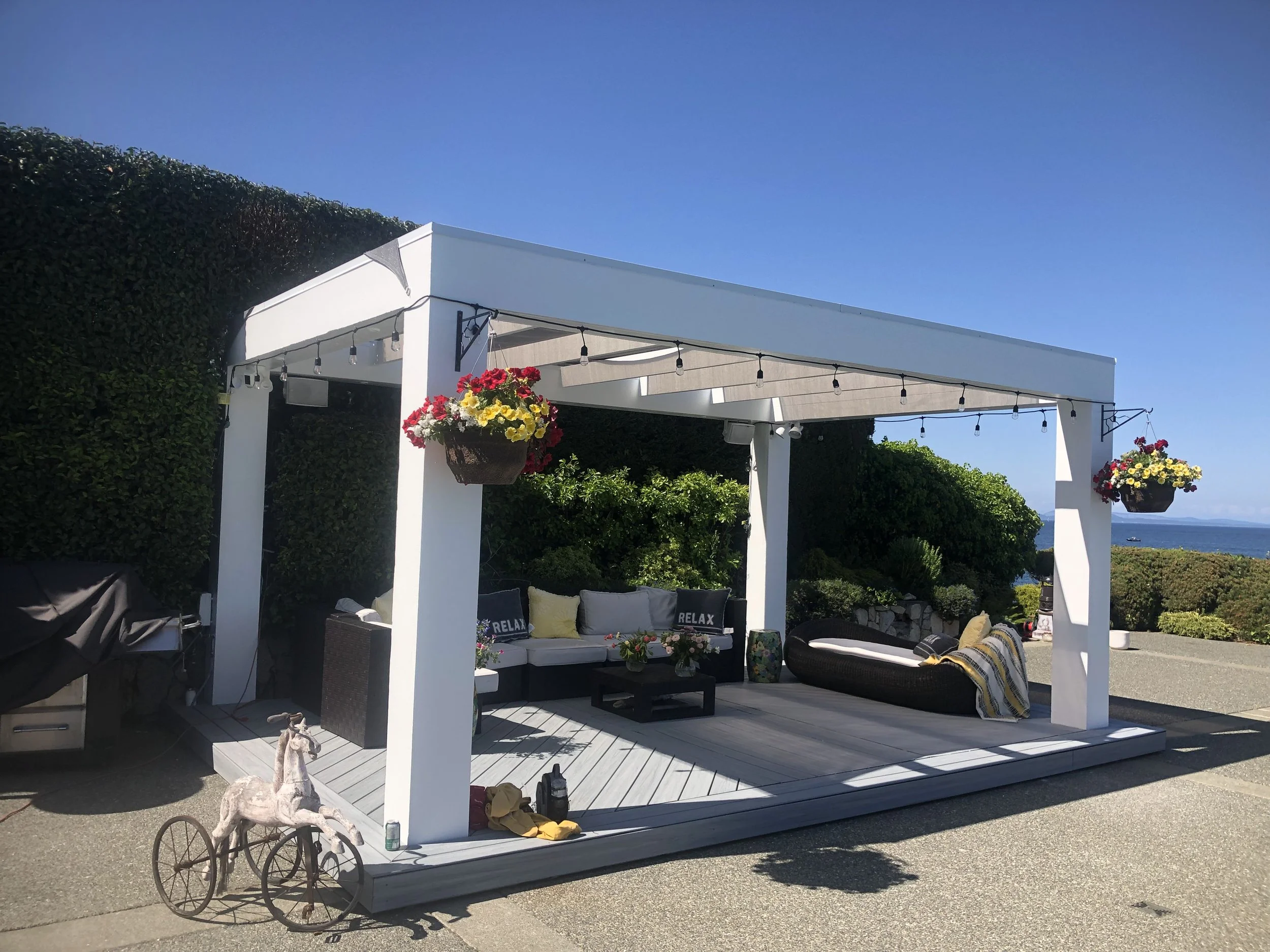 Outdoor white pergola with string lights, hanging flower baskets, and outdoor furniture including cushions with the word 'RELAX' on them, overlooking a body of water.