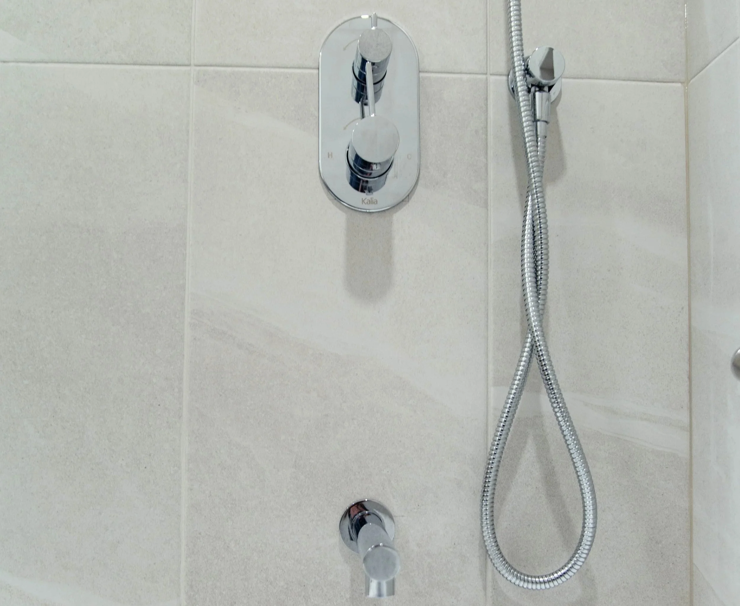 Close-up of a modern shower fixture on a tiled bathroom wall, featuring a vertical panel with two rotating control knobs and a flexible shower hose.