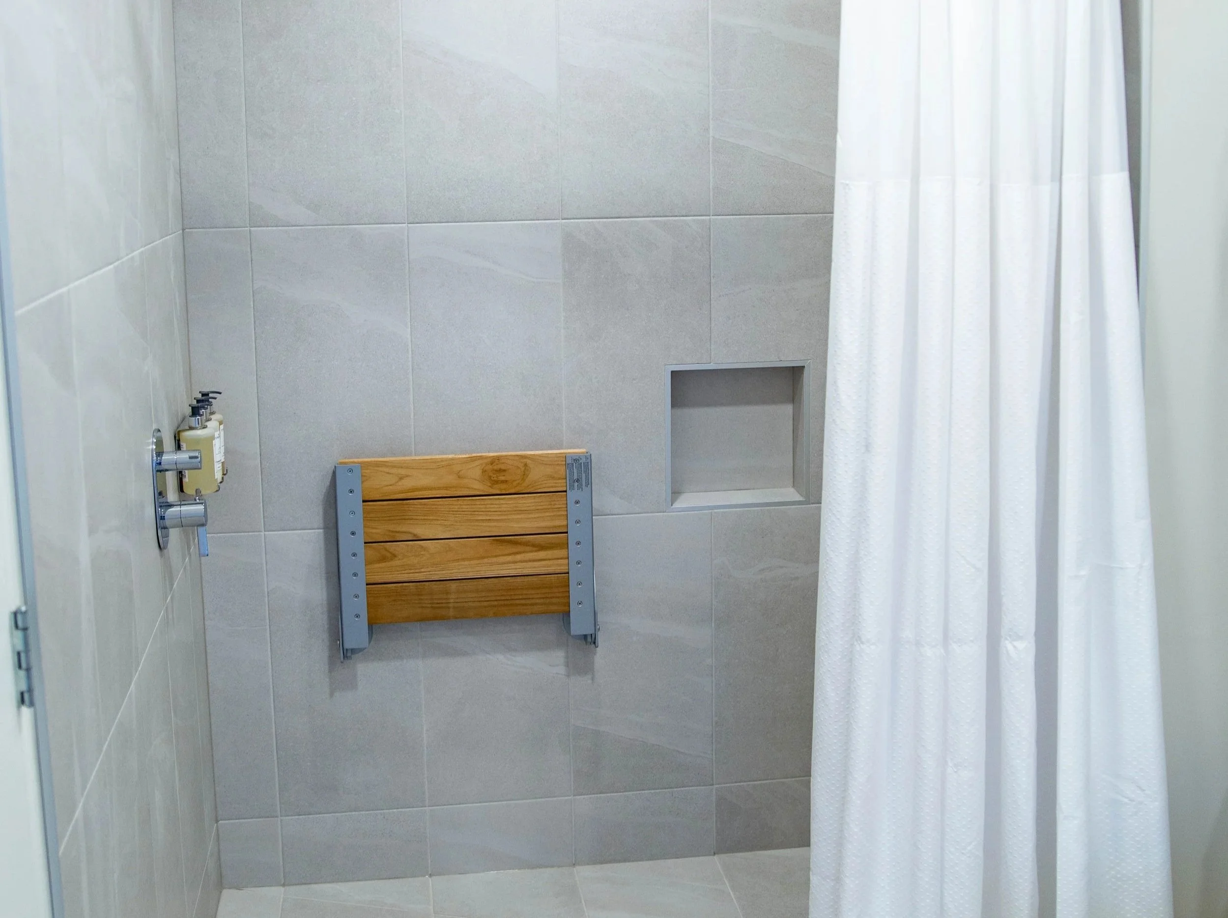 A bathroom shower area with beige tiled walls, a white shower curtain, and a wooden bench inside the shower.